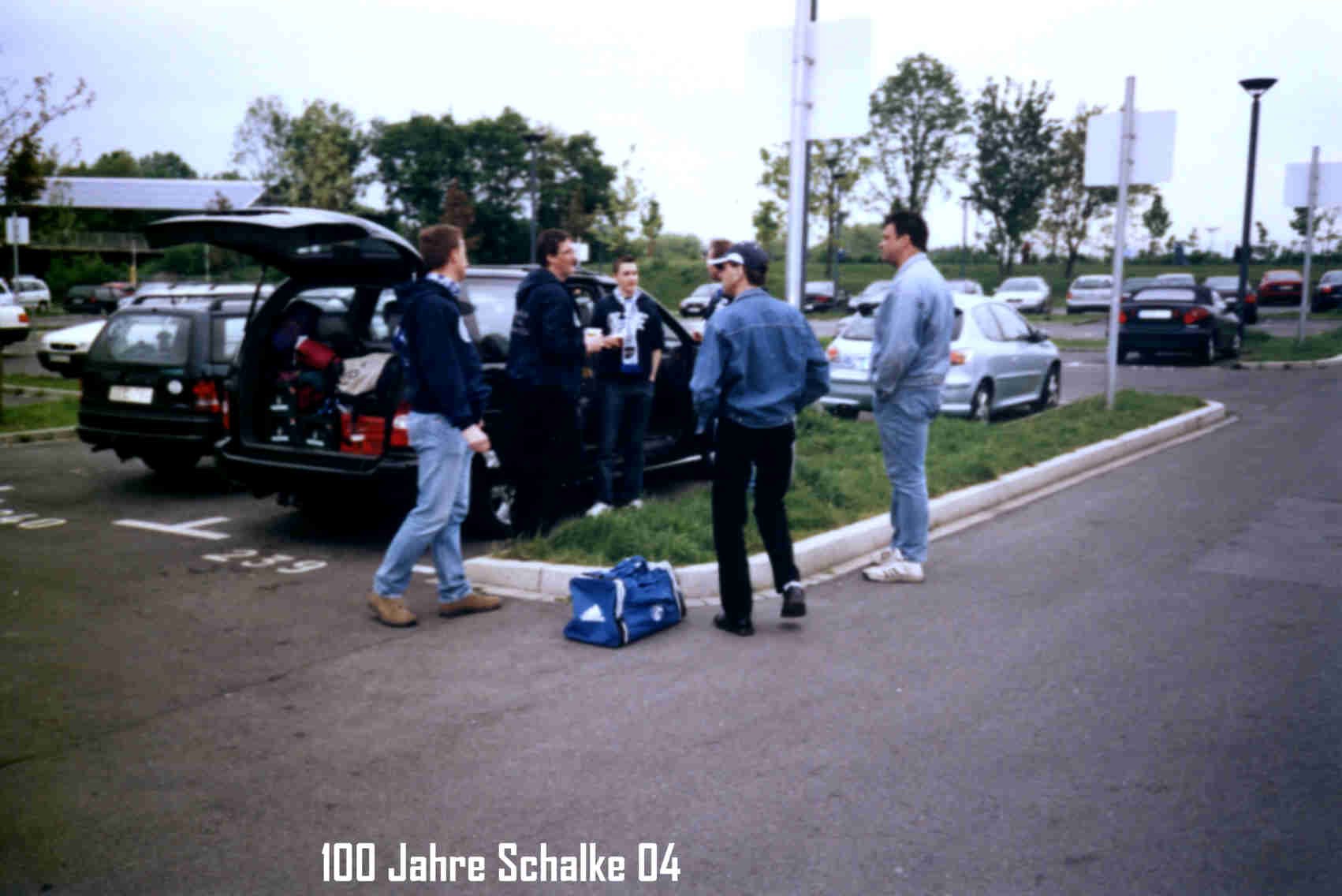 Fünf Jugendliche stehen in einem Parkplatz und unterhalten sich. Sie haben eine Tasche auf dem Boden. Es ist bewölkt und es stehen mehrere Autos im Hintergrund.