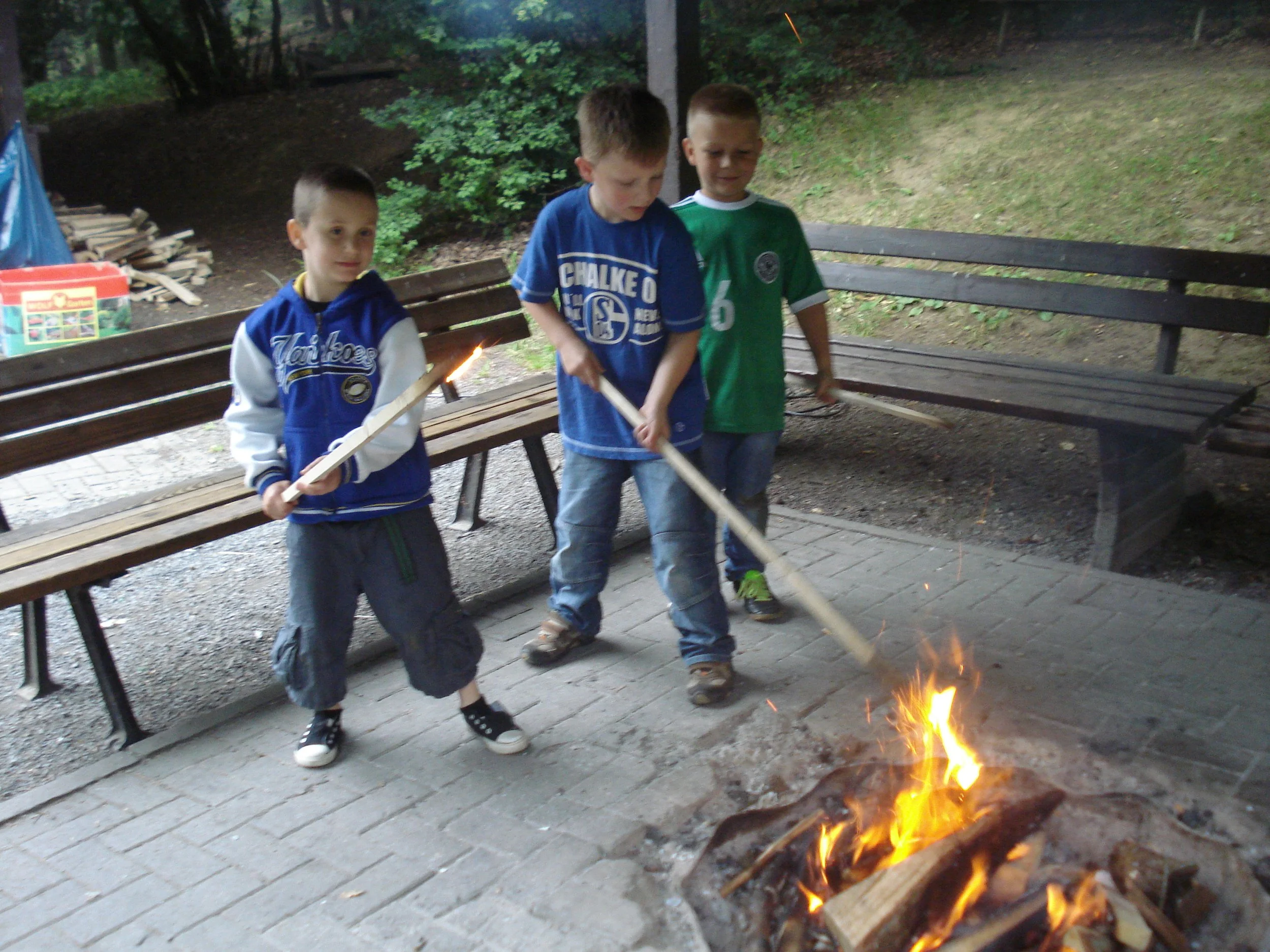 Drei Jungen stehen um ein Lagerfeuer im Freien, in einem Waldgebiet. Sie halten lange Stäbe und schauen auf das Feuer.
