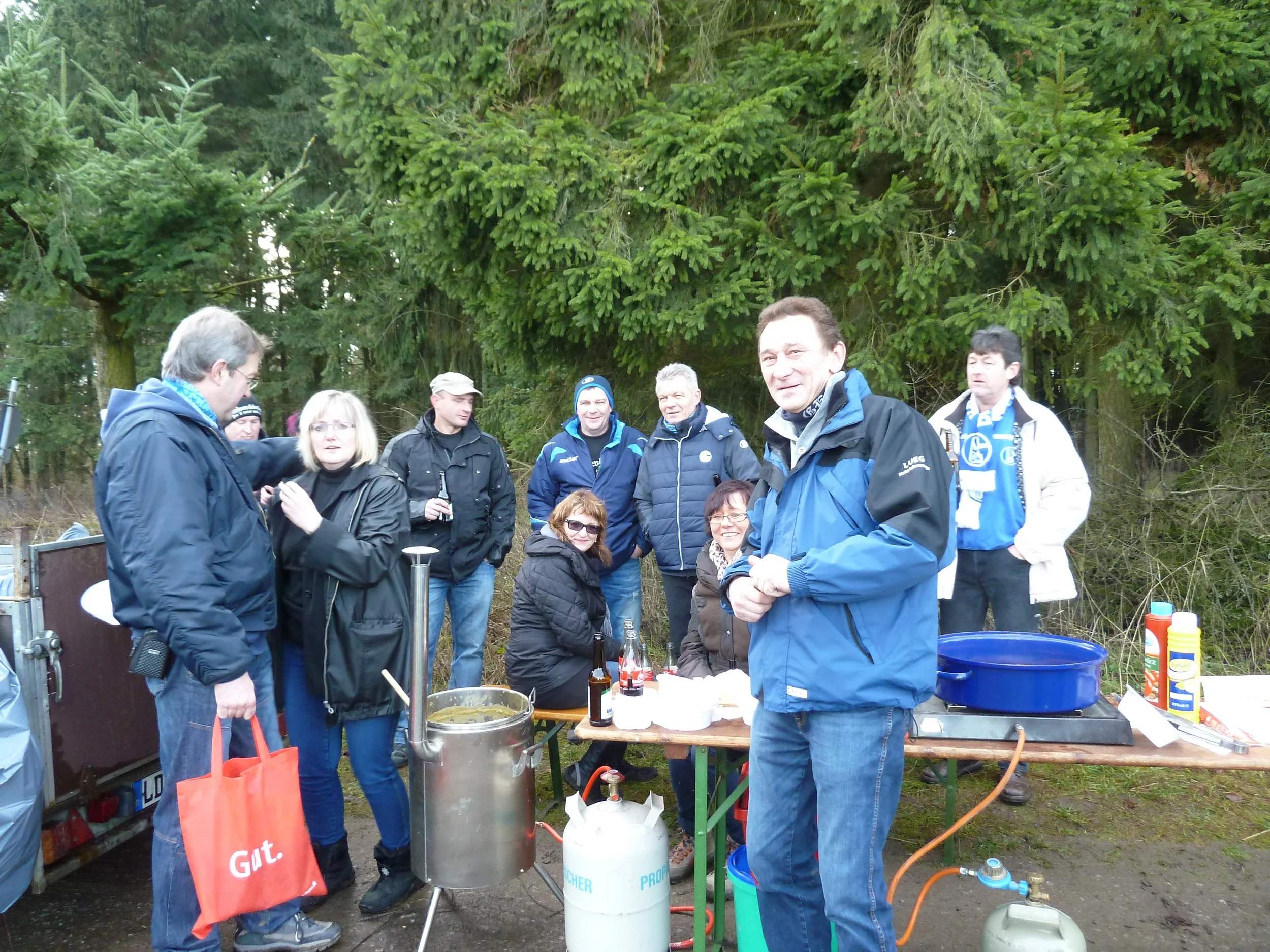 Gruppe von Menschen beim Grillen im Freien, mit Tischen und Grillstationen, umgeben von Bäumen.