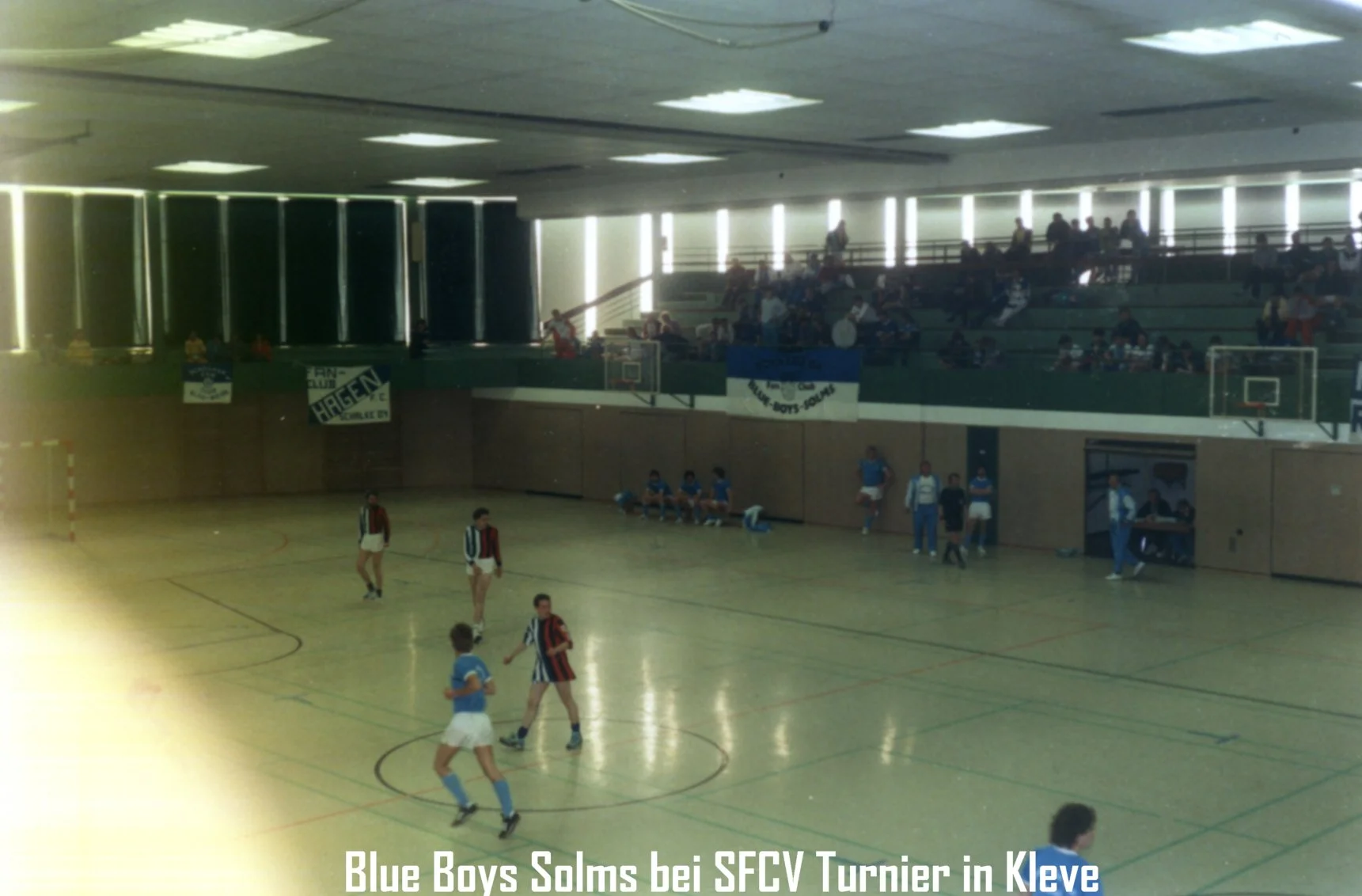 Jugendliche spielen Hallenhandball in einer Sporthalle, Zuschauer sitzen auf der Zuschauertribüne. Es gibt Handballtore und Bänke auf dem Spielfeld. Beschilderung und Banner sind sichtbar.