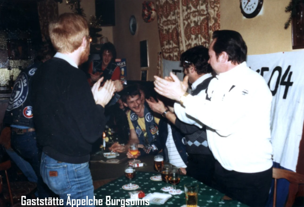Menschen in einer Bar feiern und klatschen, es gibt Stühle, Tische mit Gläsern und eine Uhr an der Wand, im Hintergrund eine Person, die laut lacht.