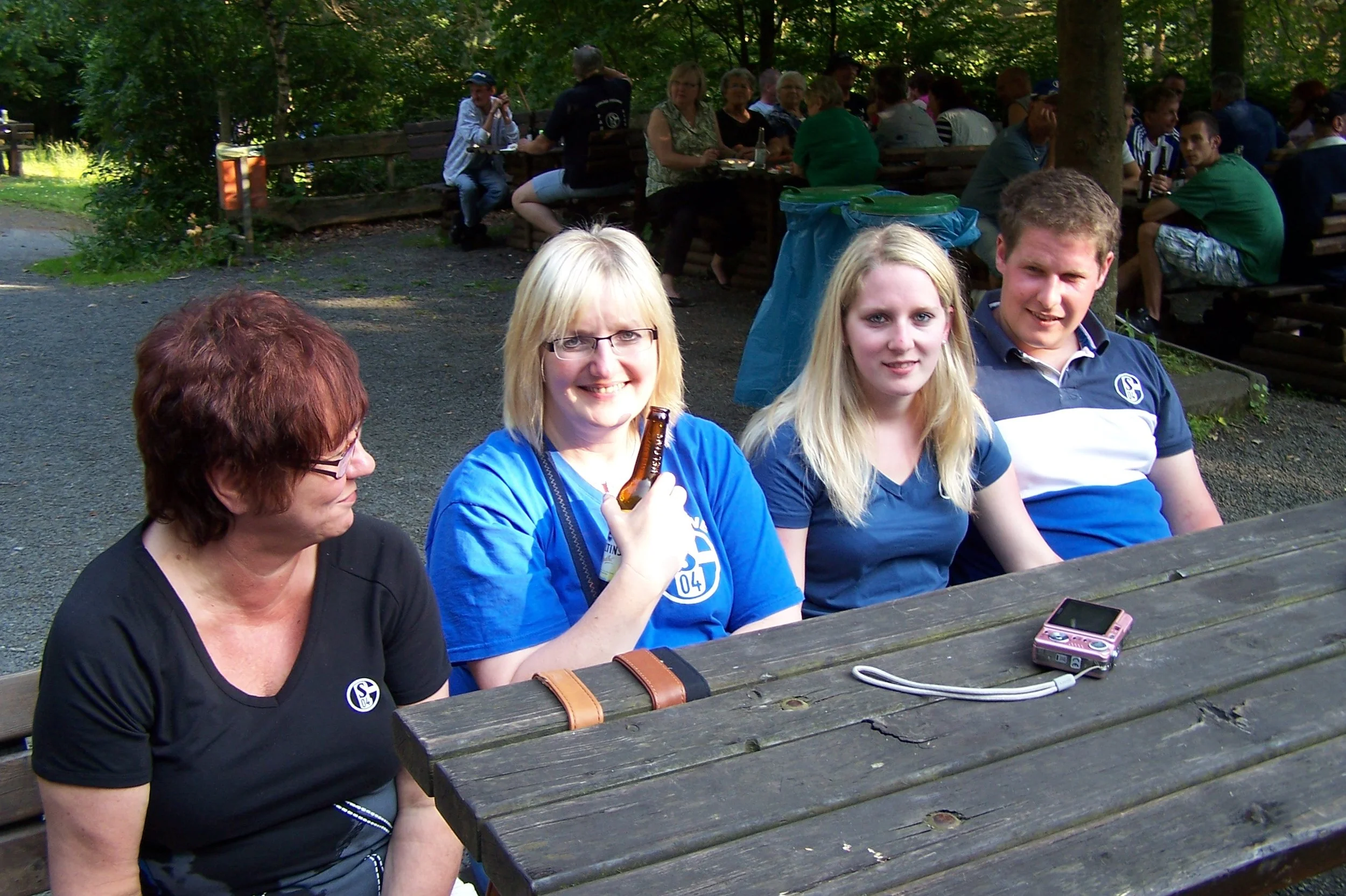 Gruppierte Menschen sitzen im Freien an Tischen unter Bäumen, einige trinken Bier, während andere sich unterhalten, es scheint eine Geselligkeit zu sein.
