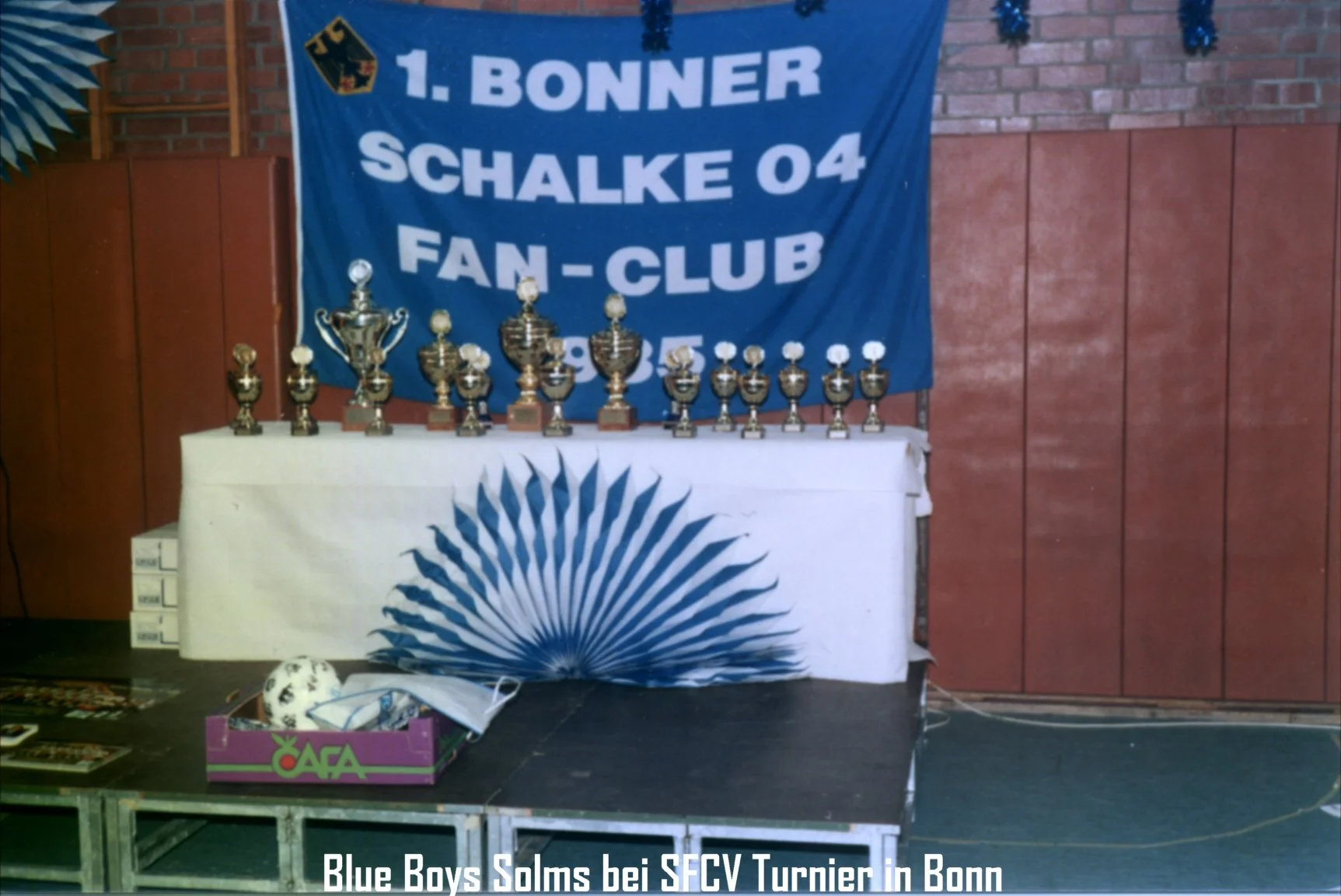 Trophäen auf einem Tisch vor einem blauen Banner mit Text und logos bei einem Turnier in Bonn. Der Banner trägt die Aufschrift "1. Bonner Schalke 04 Fan-Club". Auf dem Tisch liegen mehrere Pokale und ein Fußball. Im Vordergrund sind einzelne Gegenstä