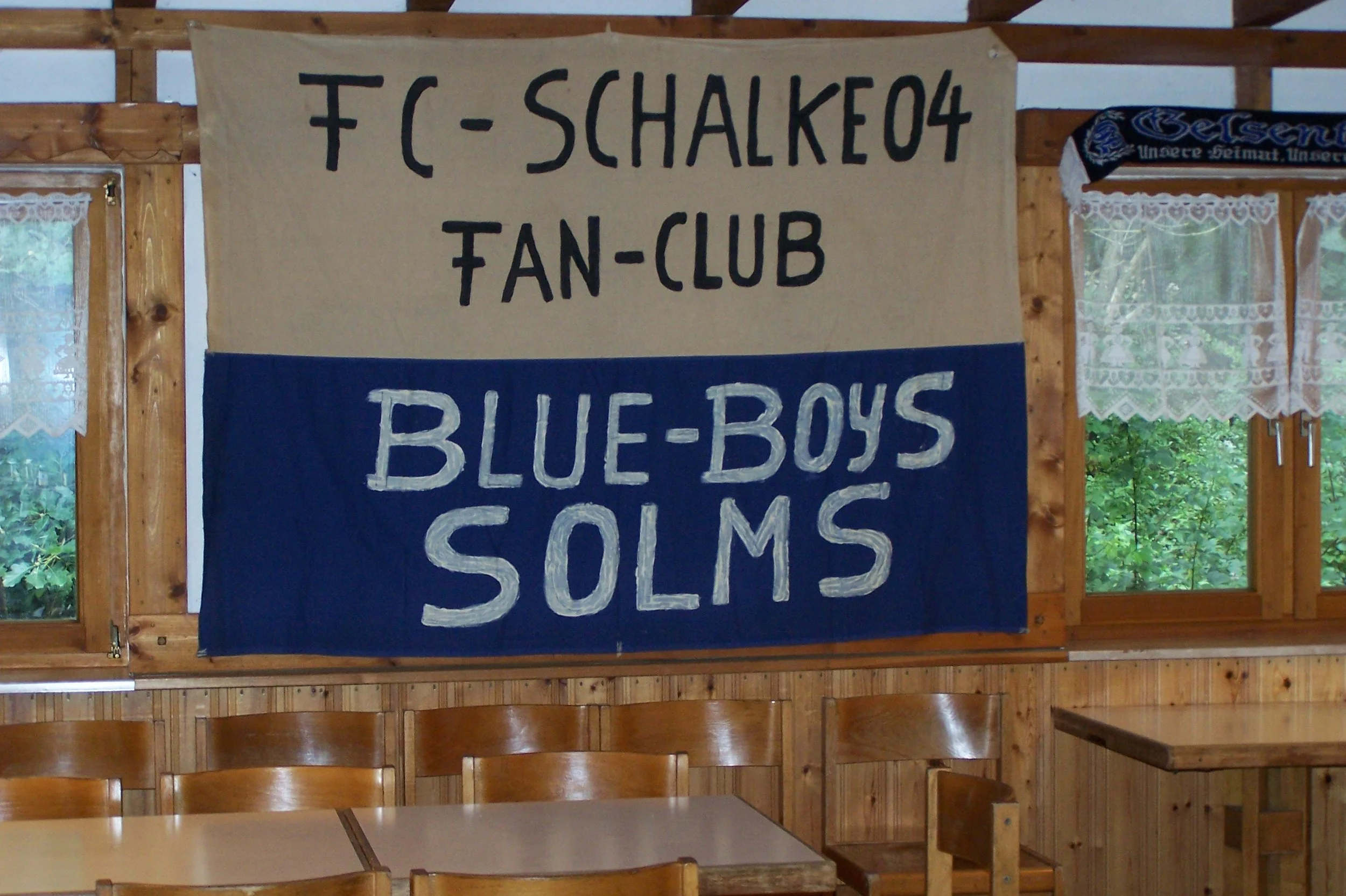 Ein Wandbild mit den Aufschriften 'FC Schalke 04', 'Fan-Club' und 'Blue-Box' in einer Hütte aus Holz, mit Fenstern und Holzvertäfelung.