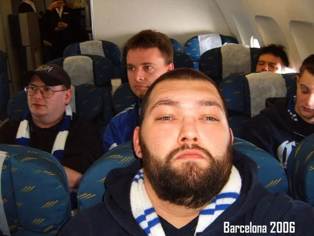 Menschen sitzen im Flugzeug, einige tragen Schals in Blau und Weiß, der Vorderste hat einen Bart, und es steht geschrieben 'Barcelona 2006' im unteren rechten Bereich.