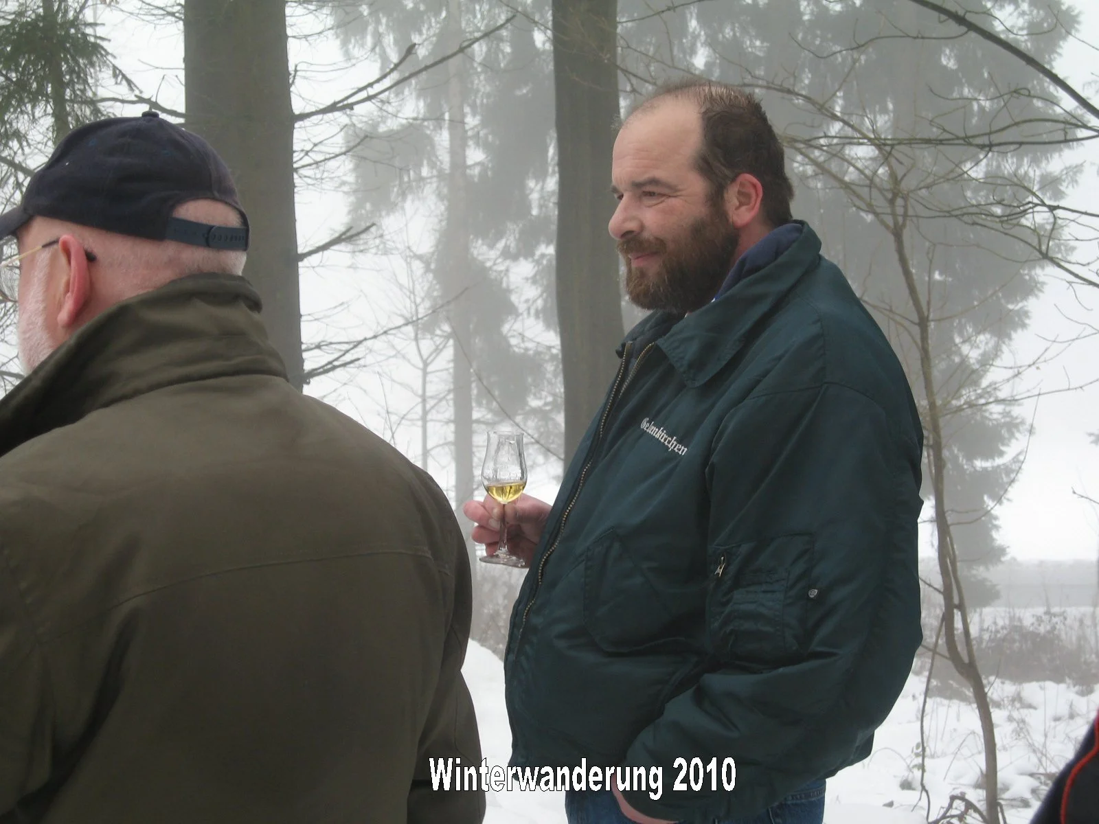 Zwei Männer bei einer Winterwanderung im verschneiten Wald, einer hält ein Glas Weißwein. Es ist neblig. Der Text 'Winterwanderung 2010' ist am unteren Rand des Bildes sichtbar.