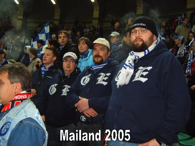 Fußballfans auf einem Stadionbesuch in Mailand 2005, viele tragen dunkle Jacken und Schal mit den Farben und Logos von Schalke 04, einer deutschen Fußballmannschaft, einige halten eine schottische Fahne, im Hintergrund sind weitere Zuschauer und Rauc