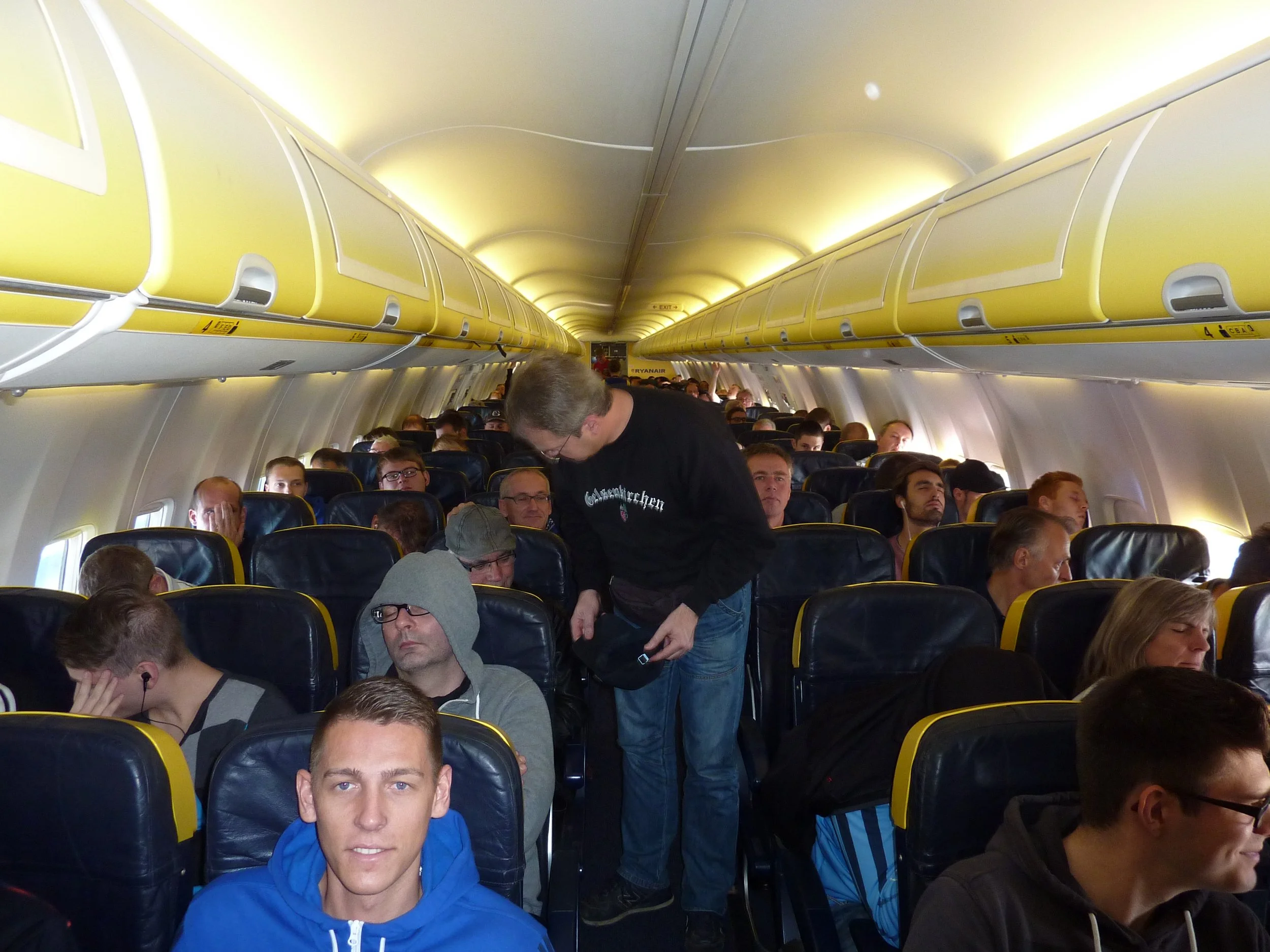 Innenaufnahme eines Flugzeugs mit mehreren Passagieren, einige sitzen, andere stehen oder bücken. Über den Sitzen sind gelbe Gepäckfächer sichtbar, und im Hintergrund ist ein Hinweis auf die Ausgänge zu sehen.