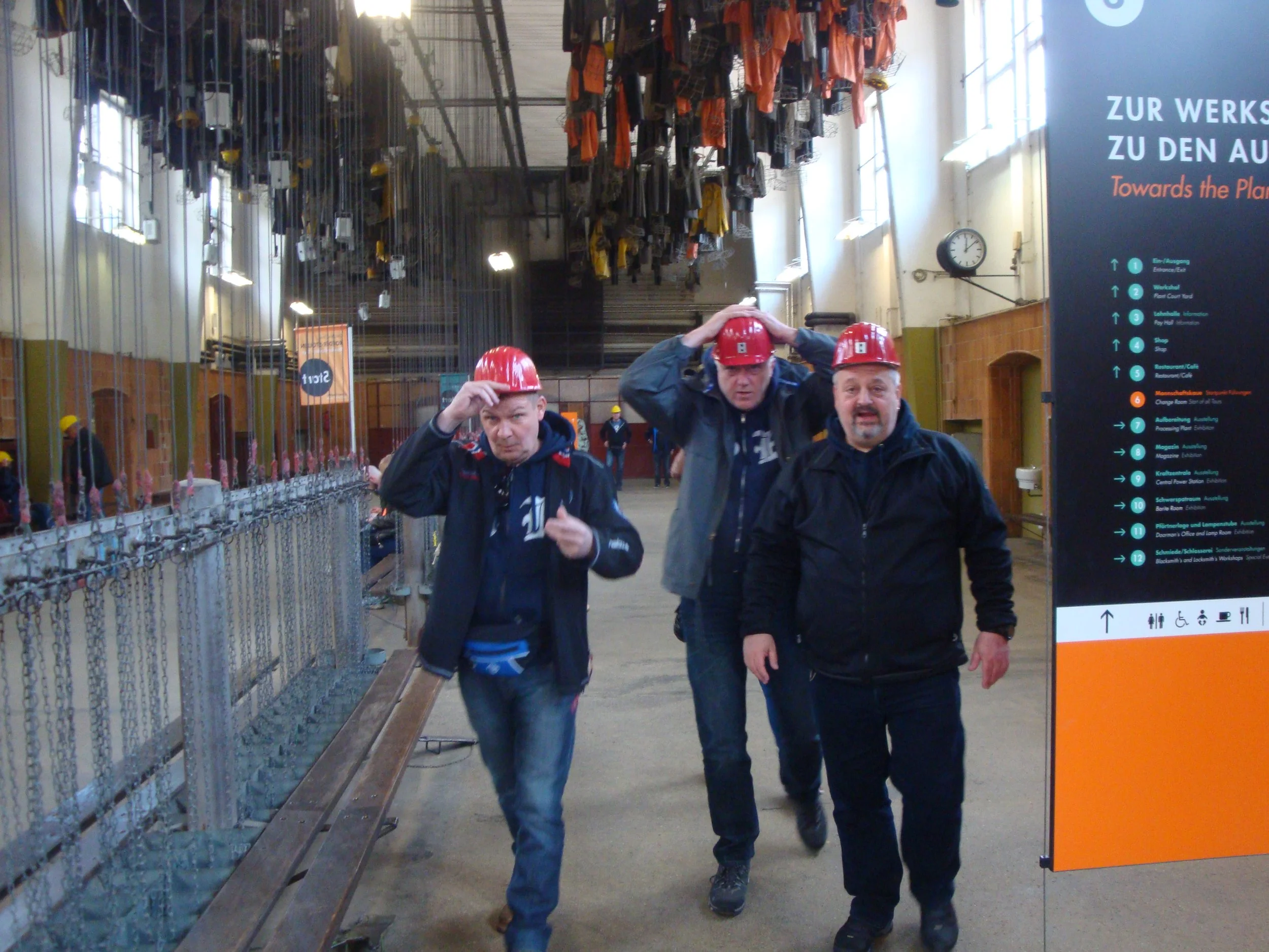 Drei Männer mit roten Helmen gehen in einem Industriegebäude. Im Hintergrund sind an einer Metallschiene befestigte Gegenstände und eine Beschilderung mit Wegweisern.