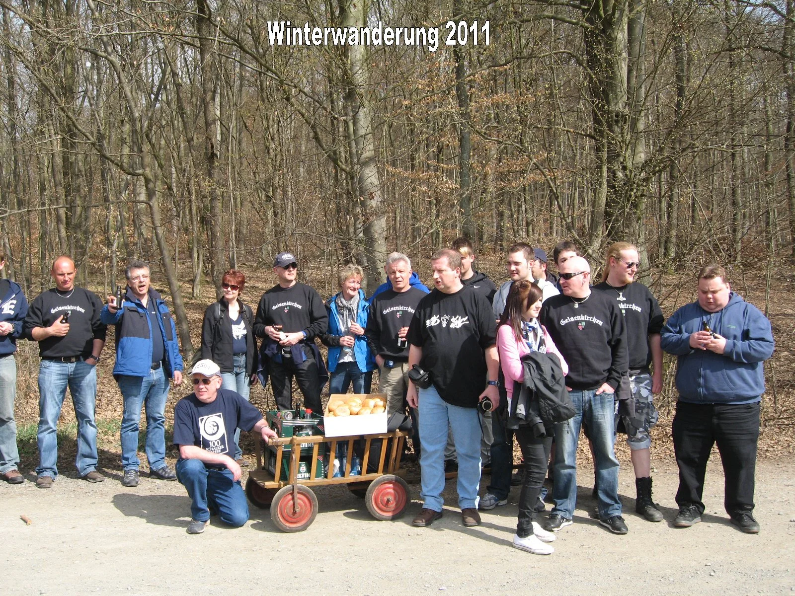 Gruppe von Menschen, die im Wald an einer Winterwanderung im Jahr 2011 teilnehmen, einige halten Getränke, es gibt eine Kiste mit Brötchen und eine kleine handkarrenartige Vorrichtung.