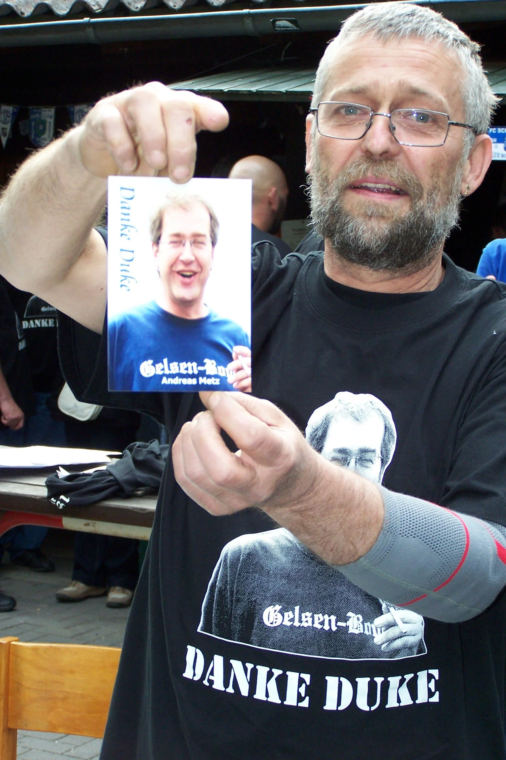 Ein Mann mit Brille und grauem Bart hält ein Foto eines anderen Mannes hoch, der lächelnd in die Kamera schaut. Beide tragen schwarze T-Shirts mit weißen Aufdrucken.
