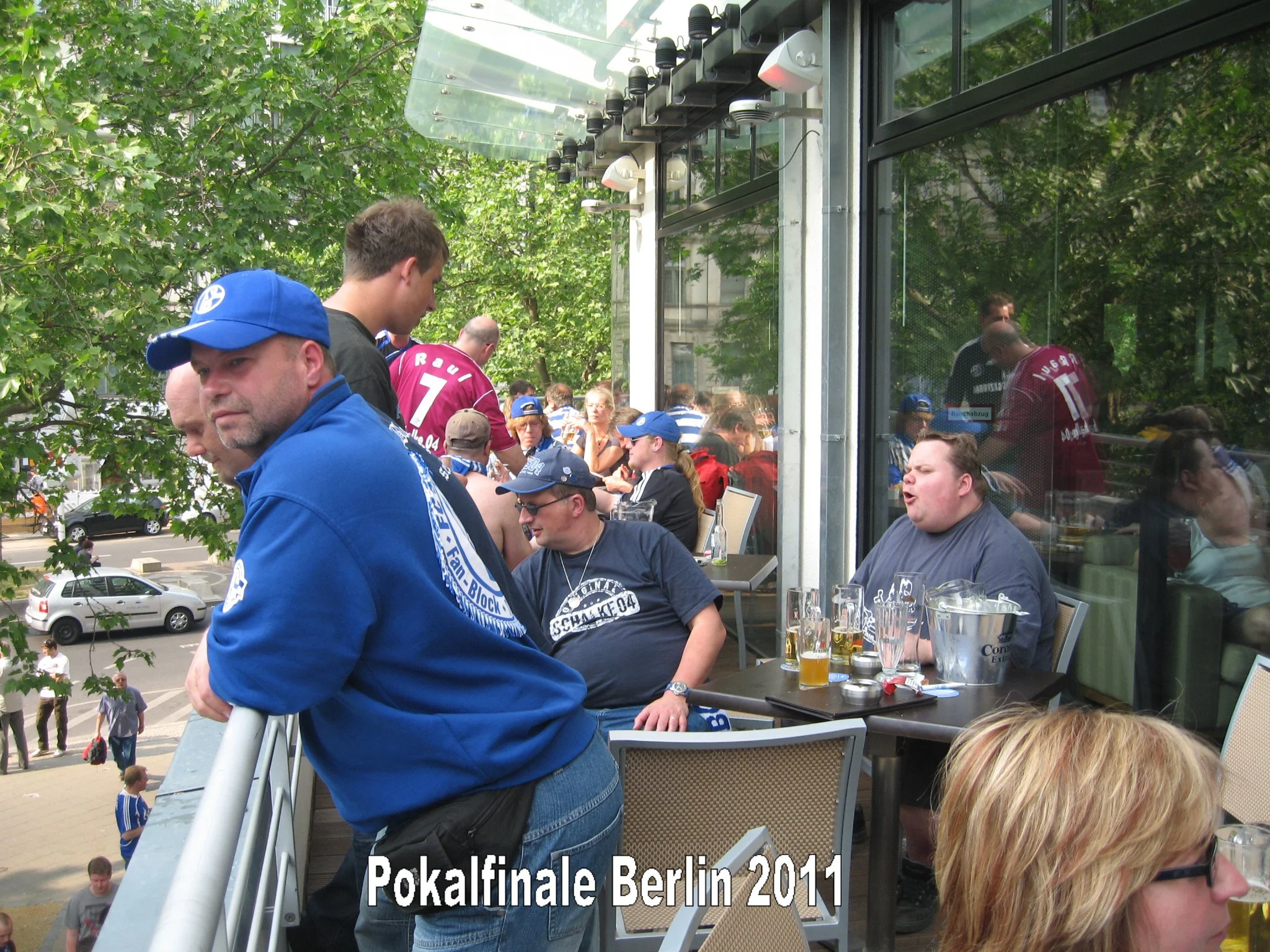 Menschen sitzen auf einer Terrasse, einige tragen Fußballtrikots, und es gibt mehrere Biergläser auf dem Tisch, im Hintergrund sind Bäume und Gebäude sichtbar.