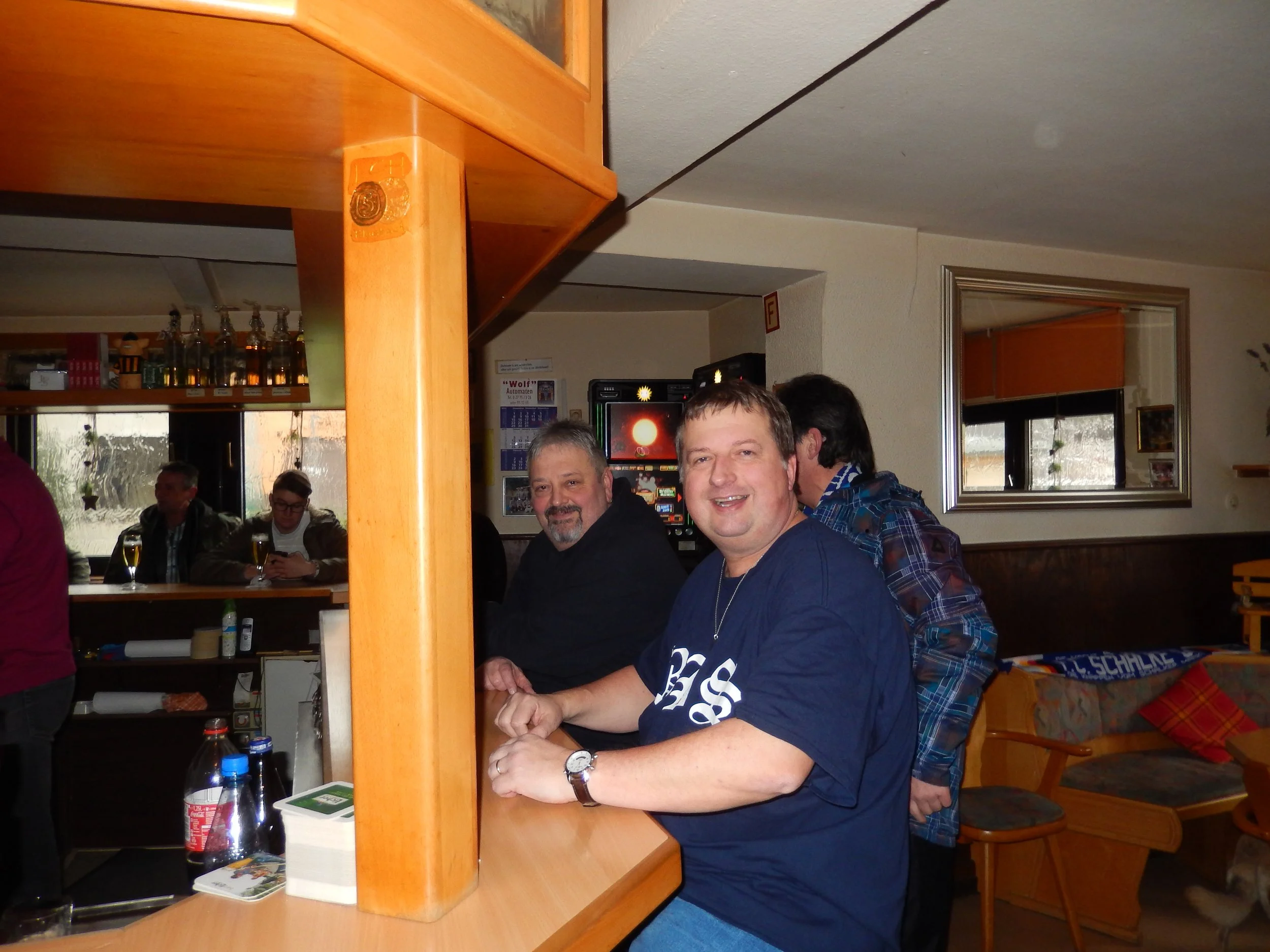 Mehrere Menschen sitzen in einer Bar oder einem Restaurant. Im Vordergrund sind zwei Männer, die an einem Tisch sitzen, während im Hintergrund andere Gäste zu sehen sind. Es gibt Getränke auf dem Tisch, und im Hintergrund sind Flaschen an der Bar sic