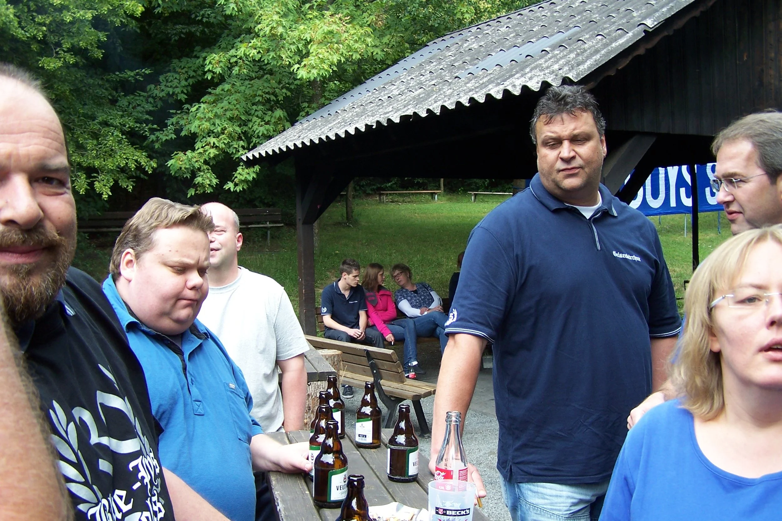 Gruppe von Menschen bei einer Outdoor-Veranstaltung, einige sitzen und einige stehen, mit Bäumen und einem Unterstand im Hintergrund.