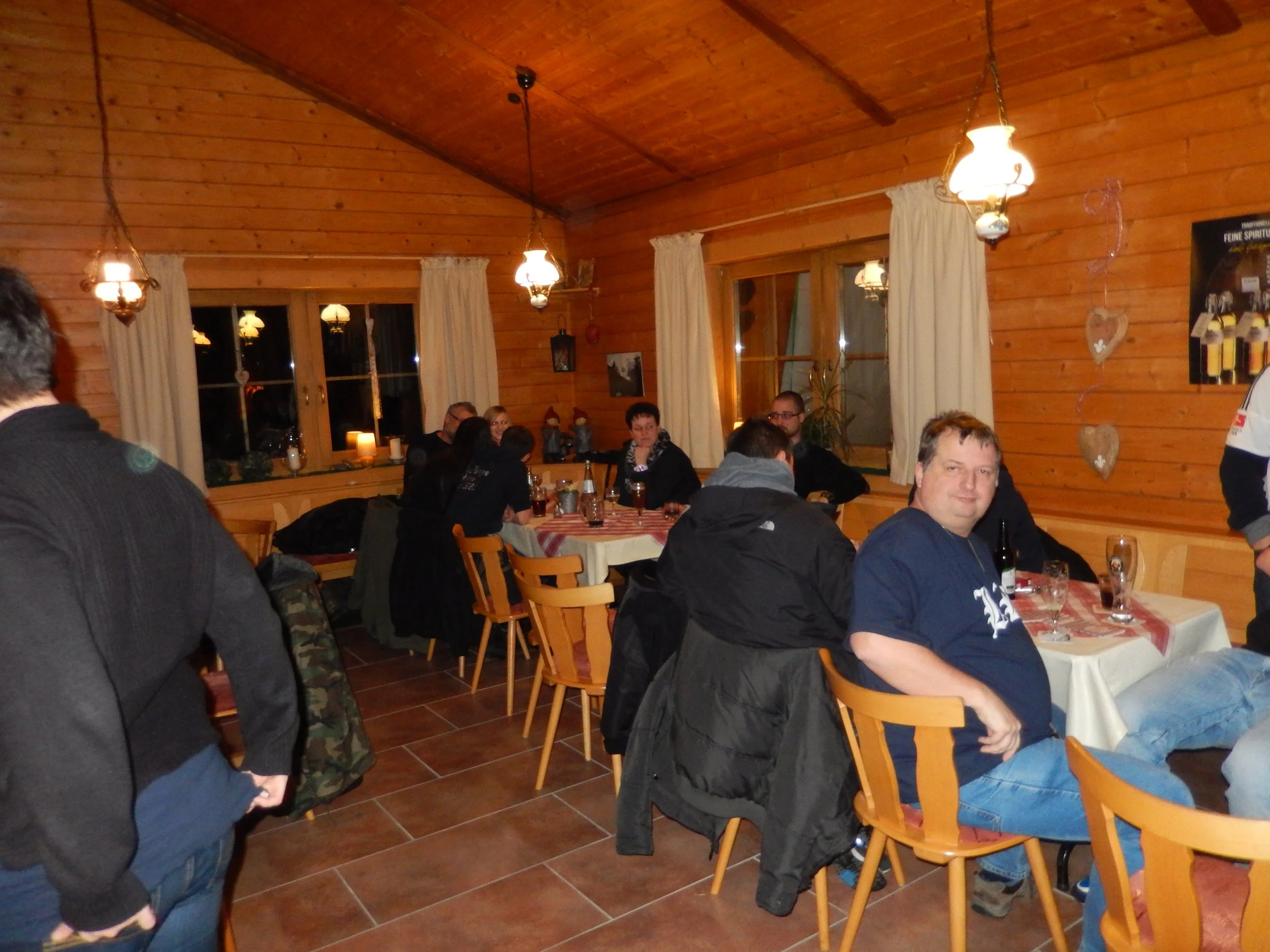 Menschen sitzen in einem rustikalen Holzraum bei einem Abendessen, einige schauen in die Kamera, andere unterhalten sich. Die Wände sind mit Holz verkleidet, und es hängen Lampen und Dekorationen. Es sind mehrere Tische mit gedeckten Tischen vorhande