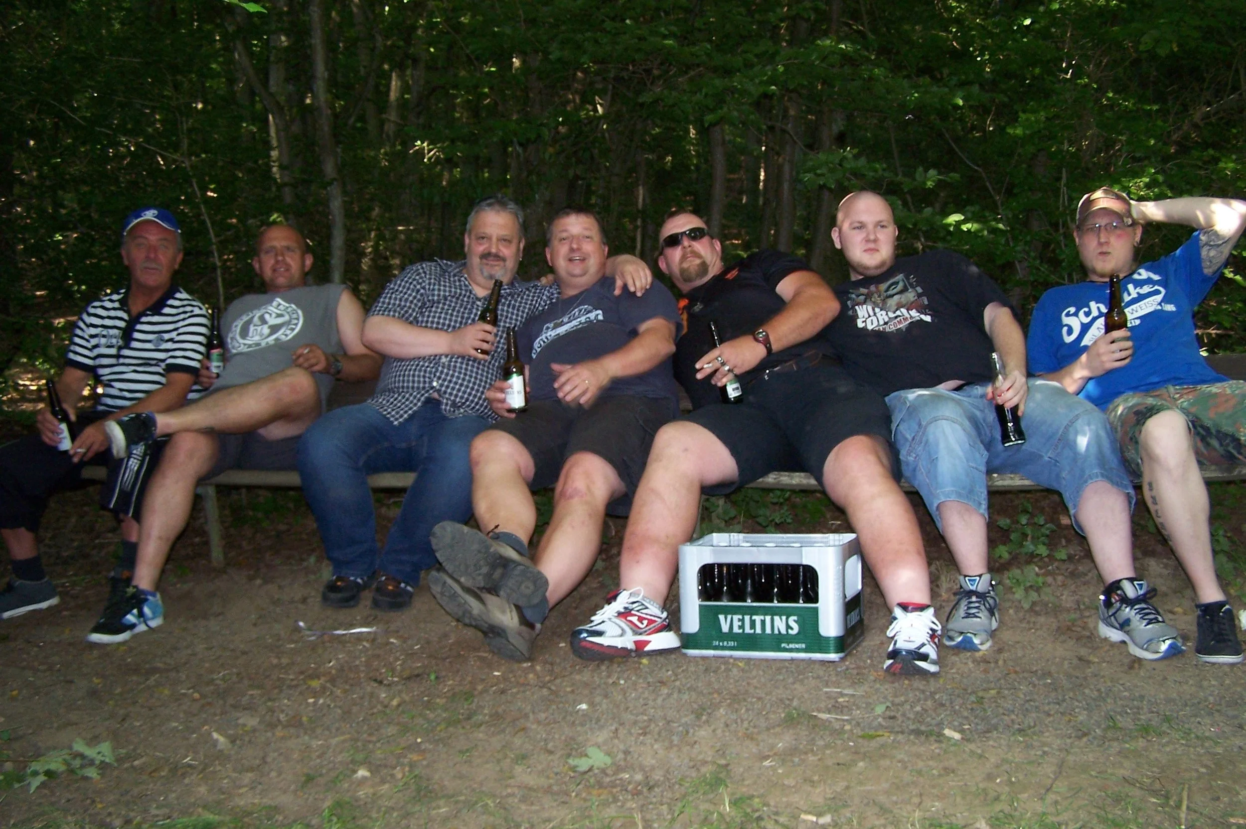 Sieben Männer sitzen auf einer Bank im Wald, halten Bierflaschen und lachen. Es sieht nach einer geselligen Zusammenkunft aus.