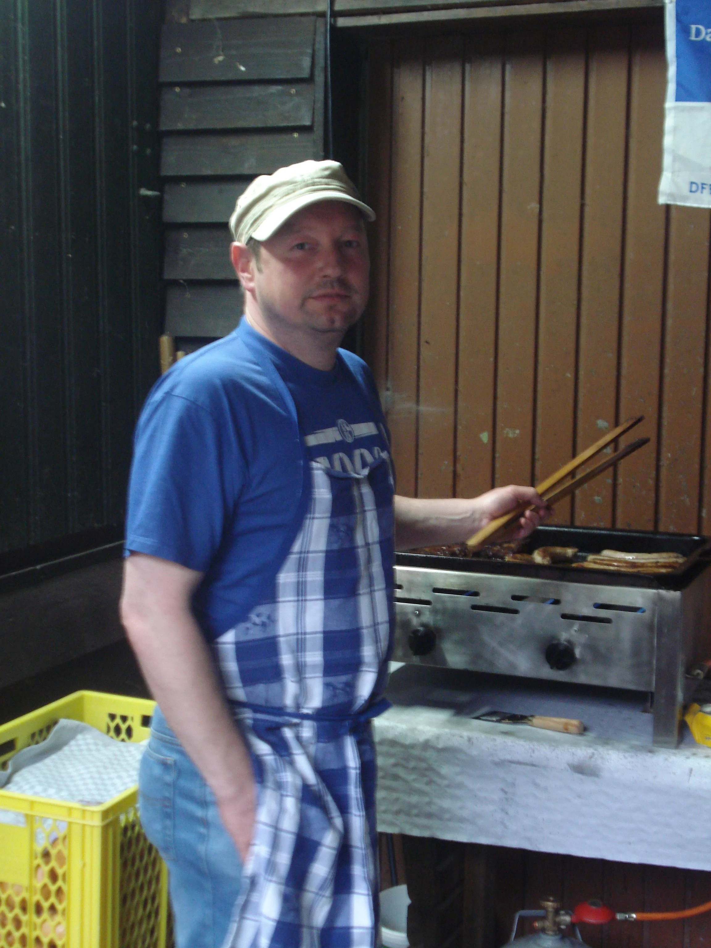 Ein Mann kocht auf einem Grill, trägt ein graues Cap und ein blau-weiß kariertes Küchenschürze.