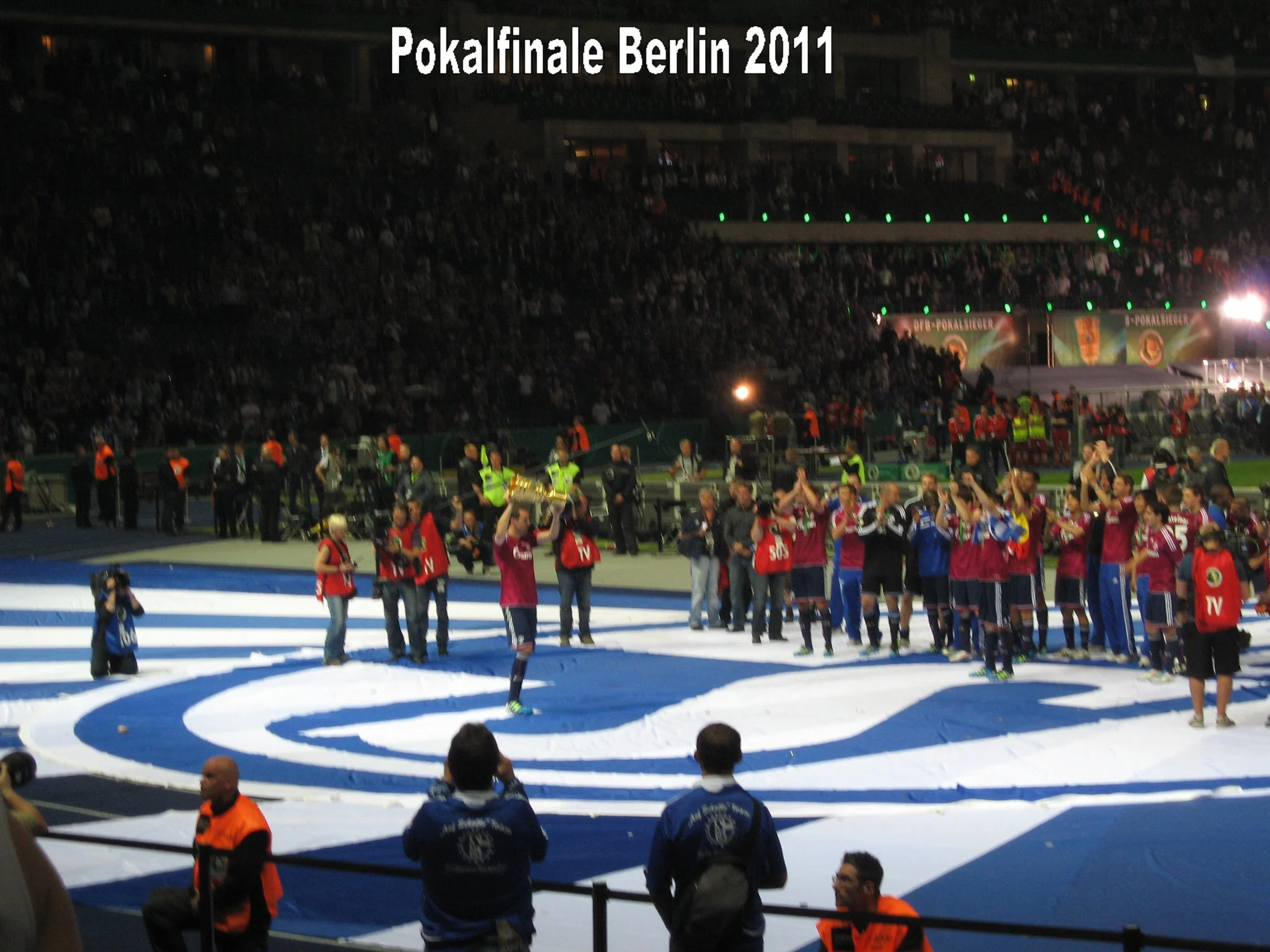 Bilder vom Finale des Pokalspiels Berlin 2011 im Fußball. Menschen feiern nach dem Spiel, einige tragen Trikots, andere halten Produkte des Vereins in den Händen. Das Stadion ist gut gefüllt mit Zuschauern.