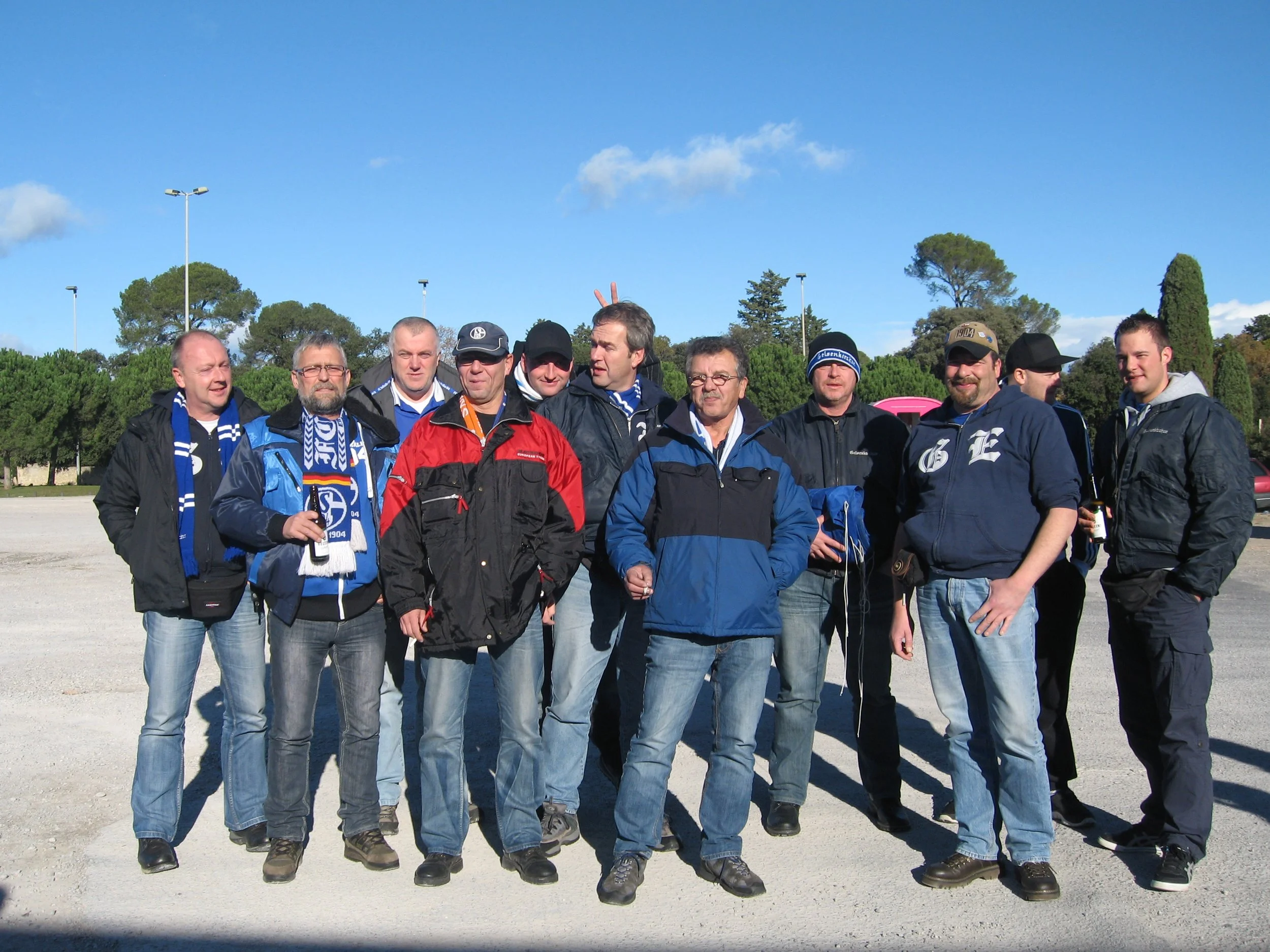 Eine Gruppe von Männern, die auf einem Platz im Freien stehen, alle tragen sportliche Kleidung und Schals, die Fußballfans repräsentieren. Im Hintergrund sind Bäume und Lichtmasten, die auf einen offenen Sportplatz oder eine Sportanlage hinweisen.