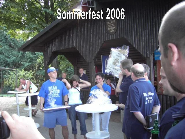 Menschen feiern Sommerfest 2006 im Freien vor einem dunklen Holzgebäude, einige stehen um Stehtische, trinke, unterhalten sich, andere schauen zu.