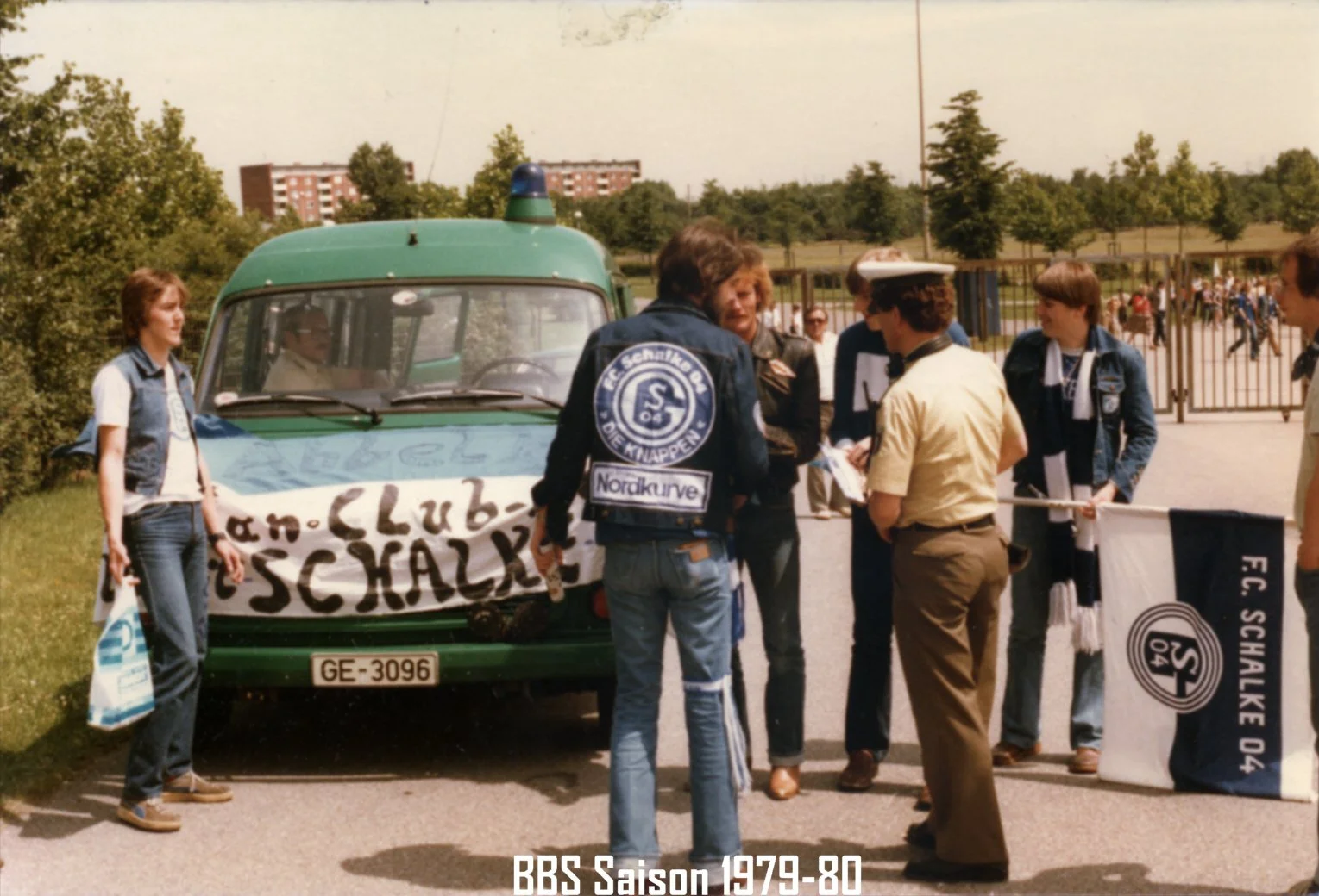 Gruppe von Menschen, die vor einem Auto mit einem Banner stehen, das den FC Schalke 04 Vereinsnamen zeigt, im Freien auf einem Parkplatz während der Saison 1979-80.