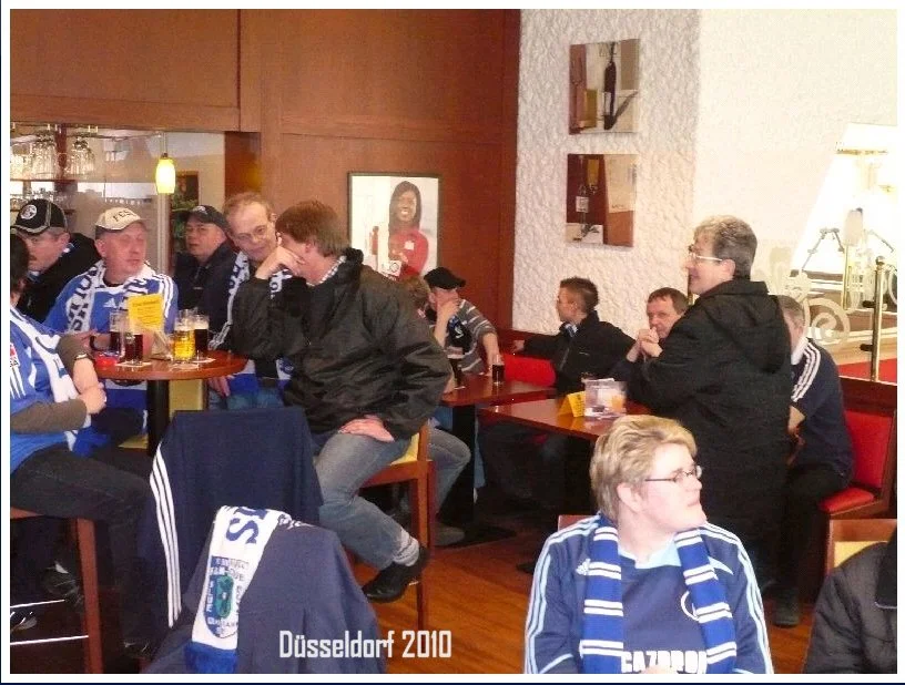 Menschen in einem Restaurant, einige tragen Sporttrikots, trinken Bier und schauen fern, im Hintergrund ein Portrait an der Wand, Text 'Düsseldorf 2010' am unteren Rand.