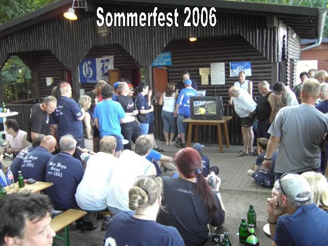 Menschen versammeln sich bei der Sommerfest 2006 Veranstaltung vor einer Holzhütte, einige sitzen an Tischen, andere stehen und schauen einen Fernseher an. Es ist tagsüber, und die Leute tragen Sommerkleidung.