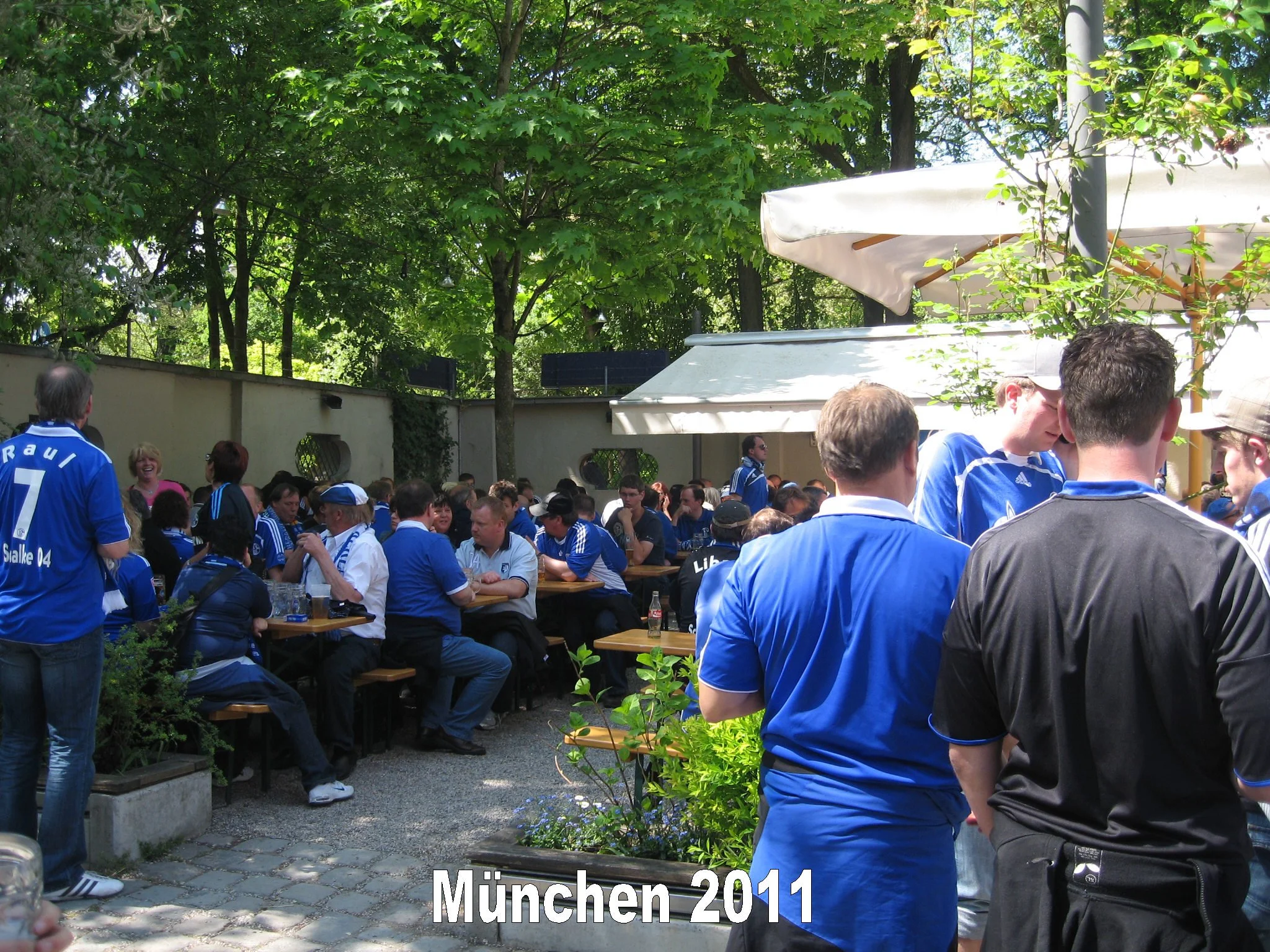 Menschen versammeln sich im Freien, einige tragen blaue Trikots, unter Bäumen mit Sonnenschutz im Hintergrund, Text: München 2011