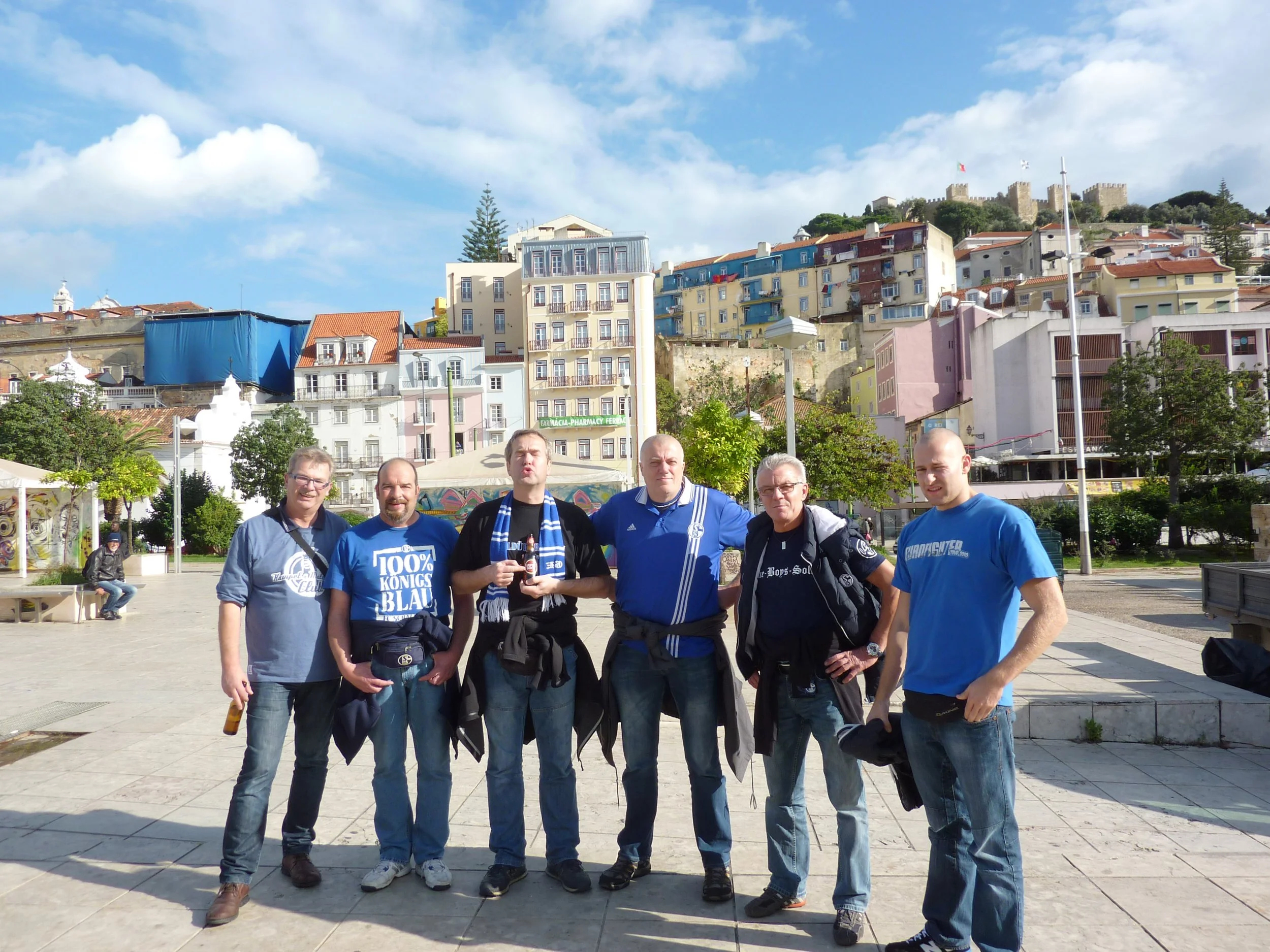 Gruppe von sechs Männern, die vor einer Stadt mit bunten Gebäuden und einer Burg auf einem Hügel stehen, bei sonnigem Himmel.