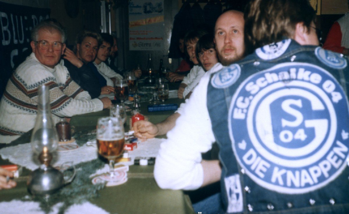 Gruppe von Menschen beim Kartenspielen in einer Bar oder einem Restaurant, einige trinken Bier, eine Person trägt eine Jacke mit ESC Schalke 04 Logo.