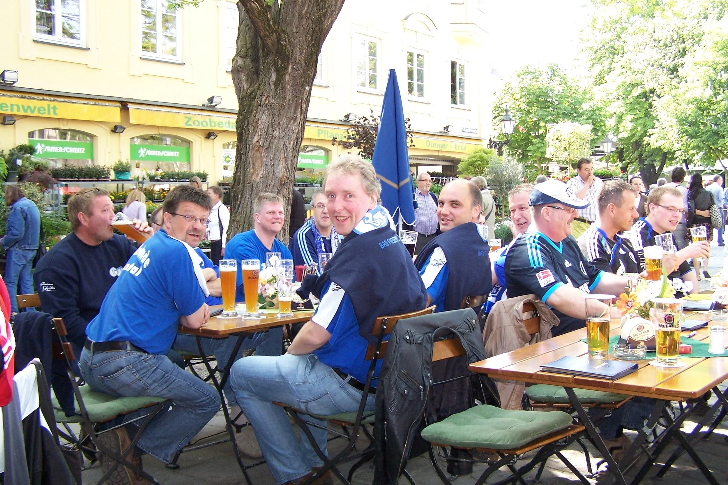 Menschen sitzen an einem langen Tisch im Freien, trinken Bier, feiern und tragen blaue Kleidung. Im Hintergrund sind Bäume, ein gelbes Gebäude mit Ladenfenstern, die mit Pflanzen dekoriert sind, sowie eine gelbe Markise mit grüner Beschriftung.