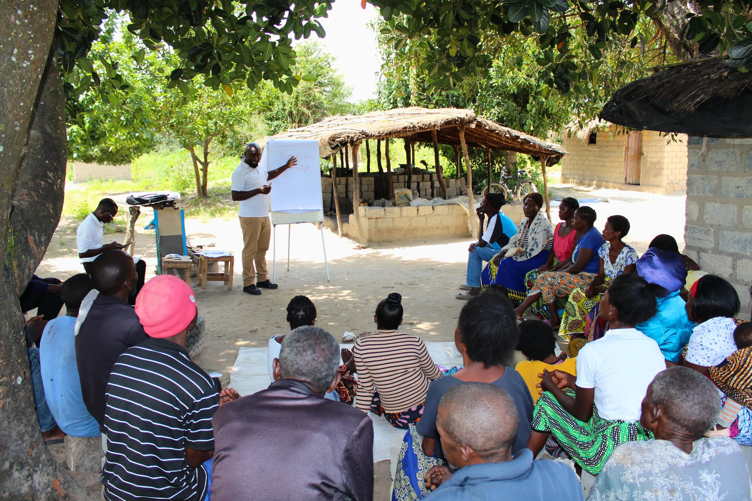 A man giving a presentation outdoors to a group of people seated under trees and a thatched roof structure. The presenter is pointing at a whiteboard with the words 'Rural Sand Filter' written on it. The setting appears to be a rural area with some buildings and bicycles in the background.