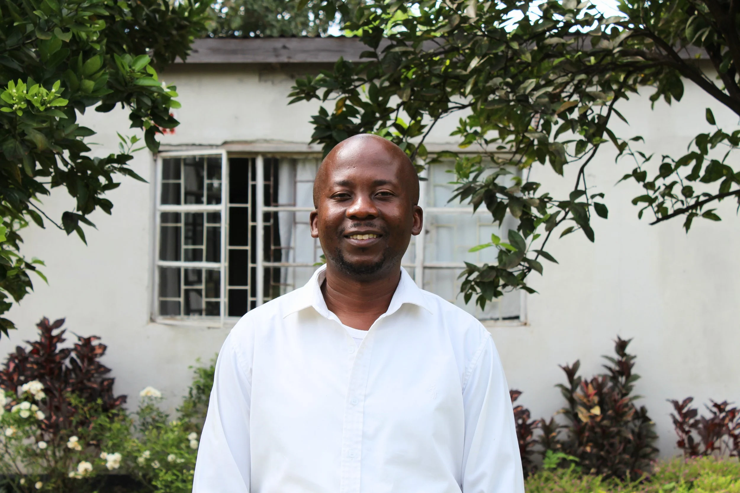 A man wearing a white shirt standing outdoors in front of a house with a window, surrounded by green bushes and plants.