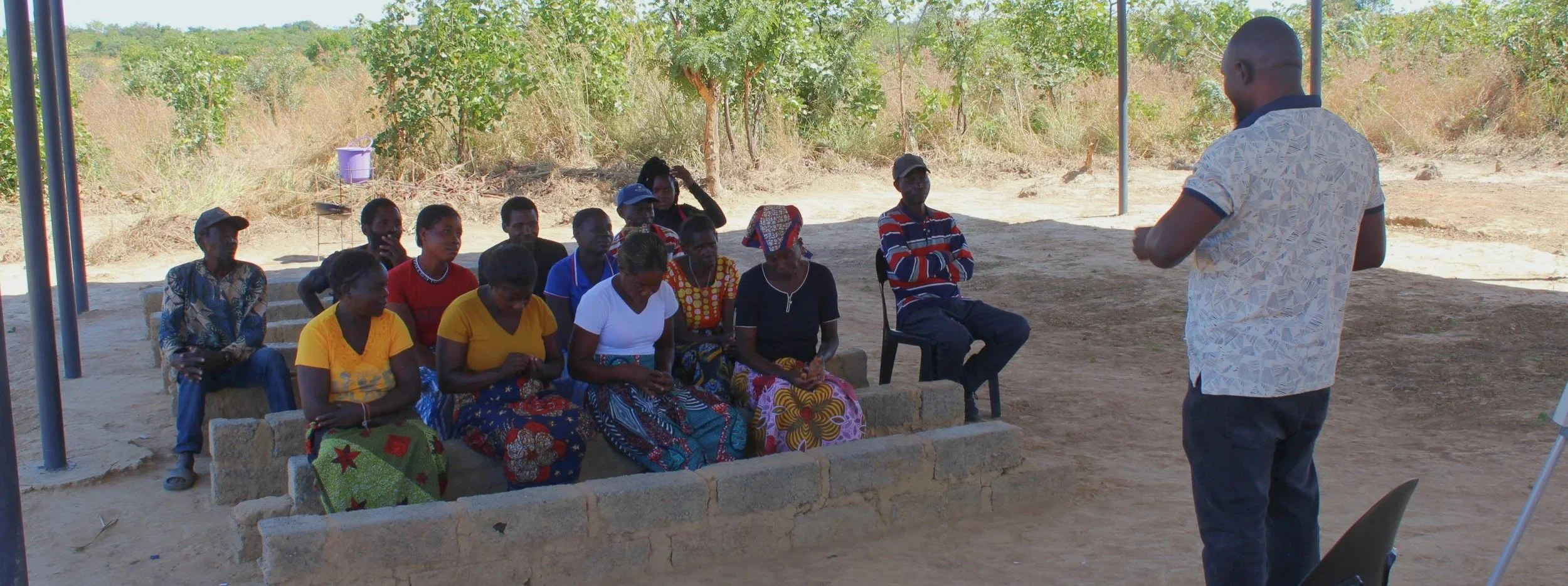 A group of people sitting outdoors on a makeshift brick and stone bench, listening to a man in a patterned shirt speaking. The setting is a natural landscape with trees and dry grass.