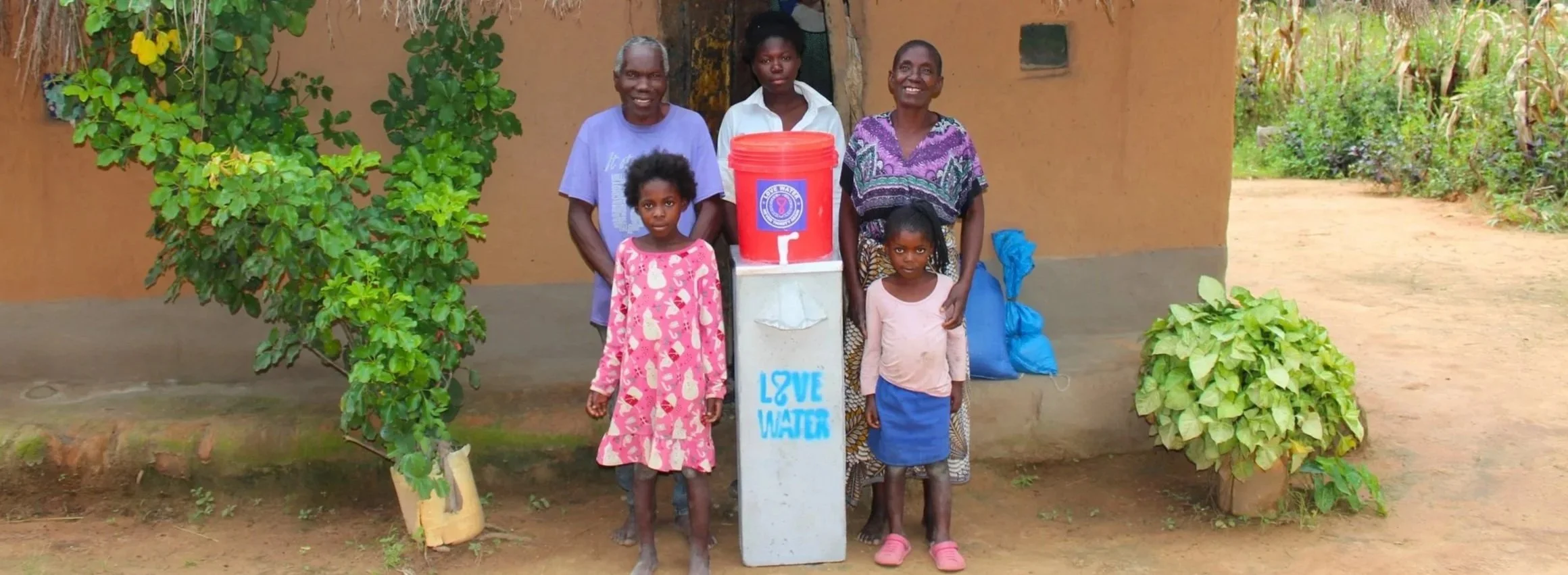 Five people, including children and adults, stand outdoors near a water dispenser with a sign that says 'LOVE WATER.' There is greenery, bushes, and a dirt pathway and building in the background.