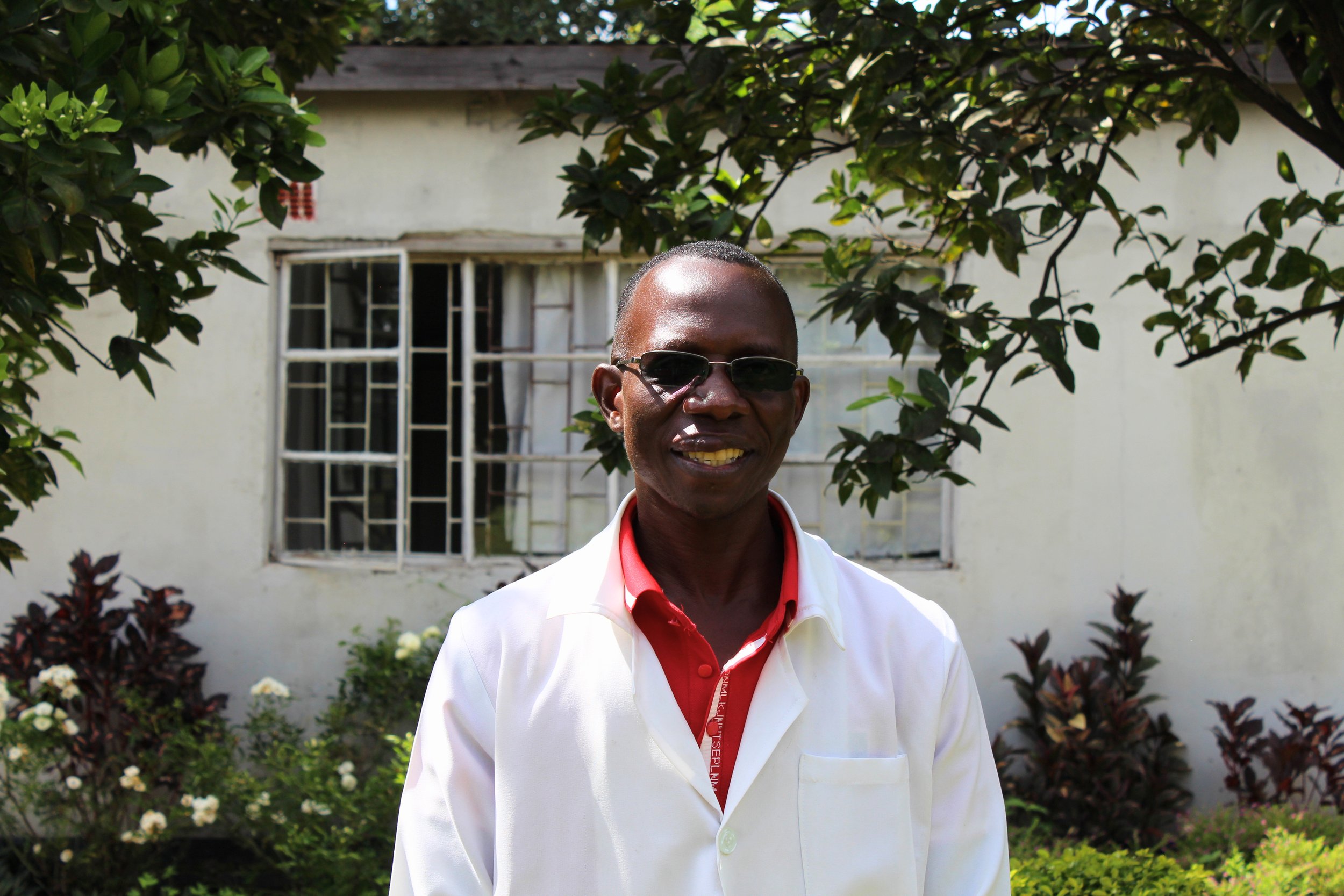 A man with dark skin wearing sunglasses and a white jacket over a red shirt stands outdoors in front of a garden with various plants and flowers, with a house in the background.