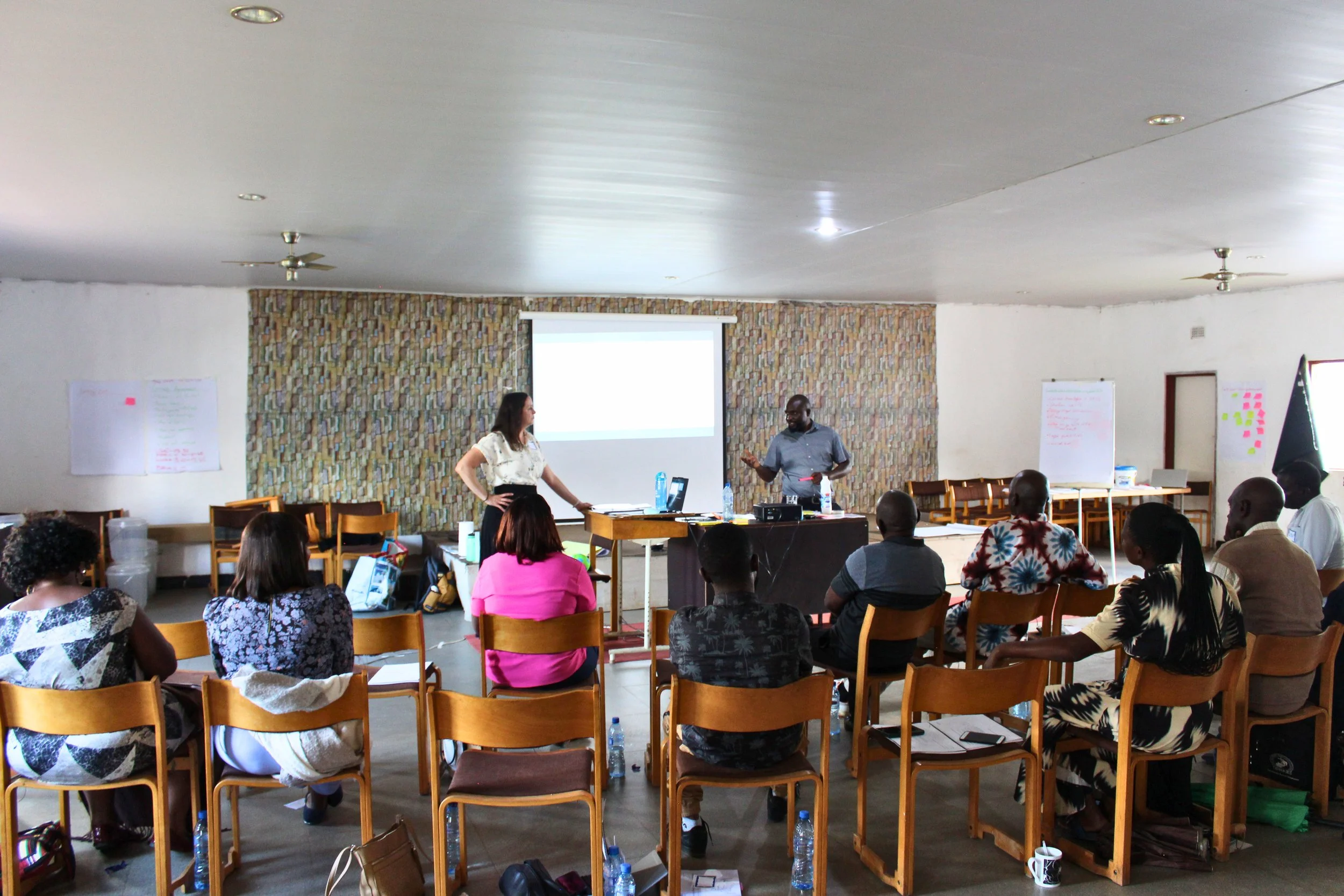 A group of people in a conference room attending a presentation. Two individuals stand at the front, one speaking and the other listening. There is a projection screen, chairs, tables, and posters with notes on the walls.