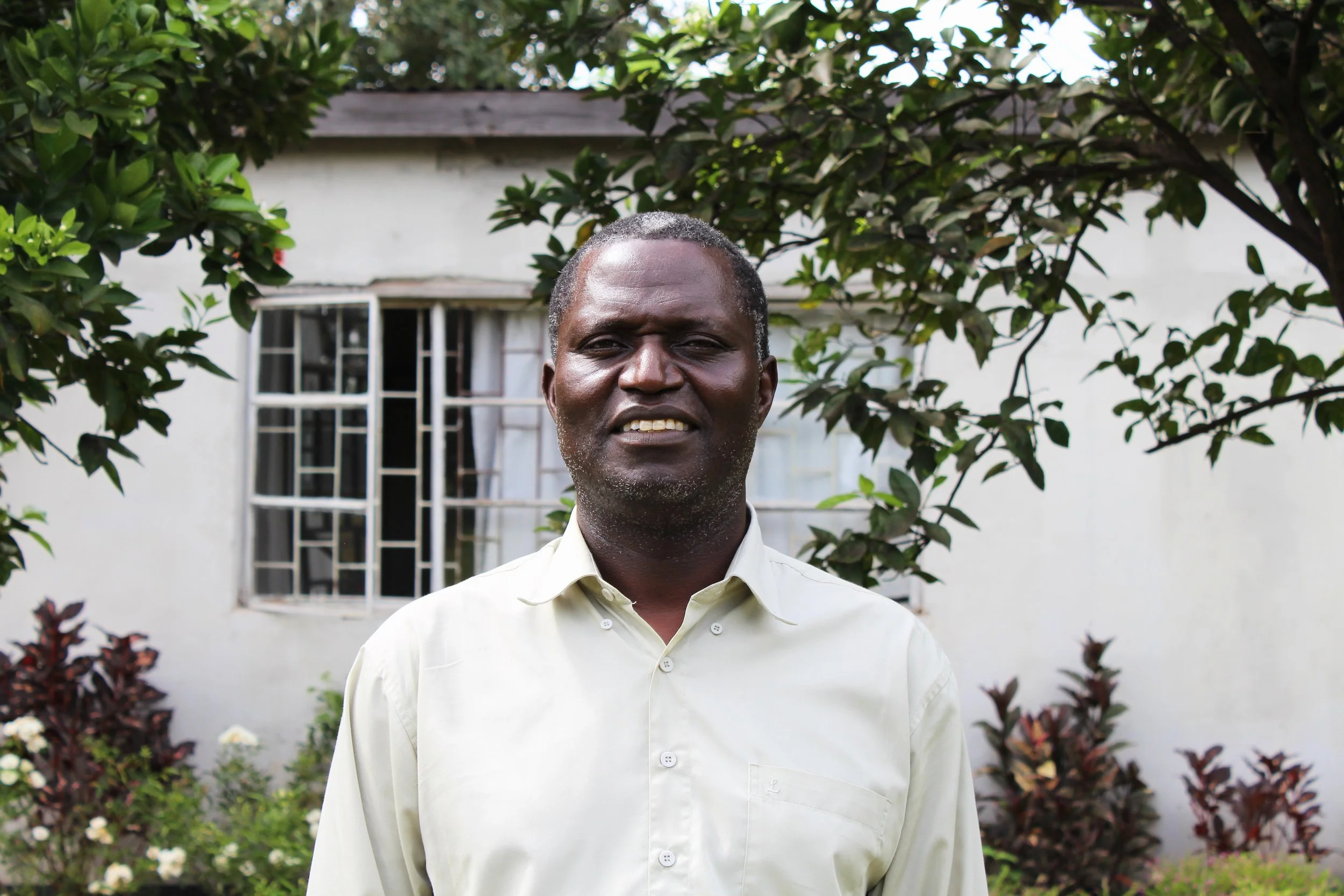 A man standing outdoors in front of a green leafy tree and a white house with barred windows, wearing a light-colored button-up shirt.