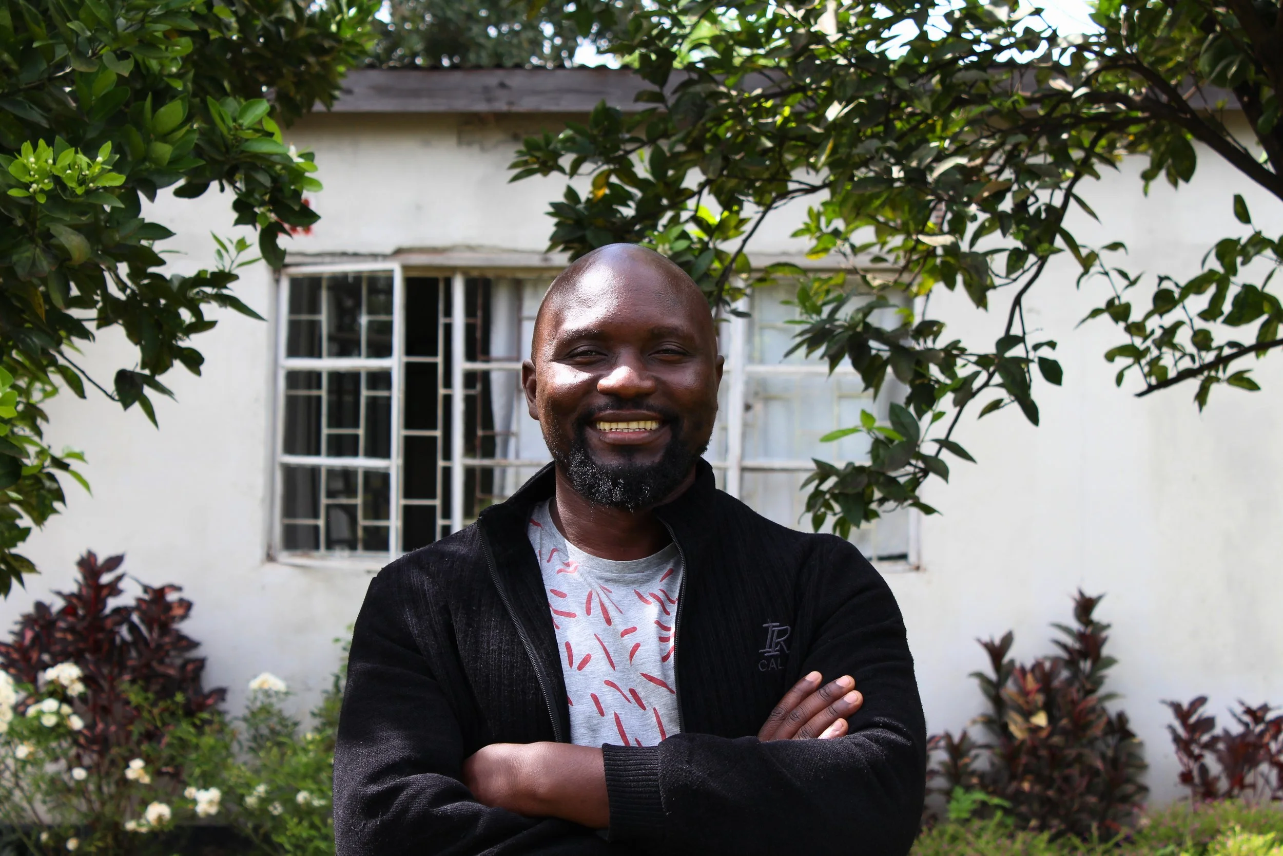 Smiling man with crossed arms standing in a garden with a white house in the background.