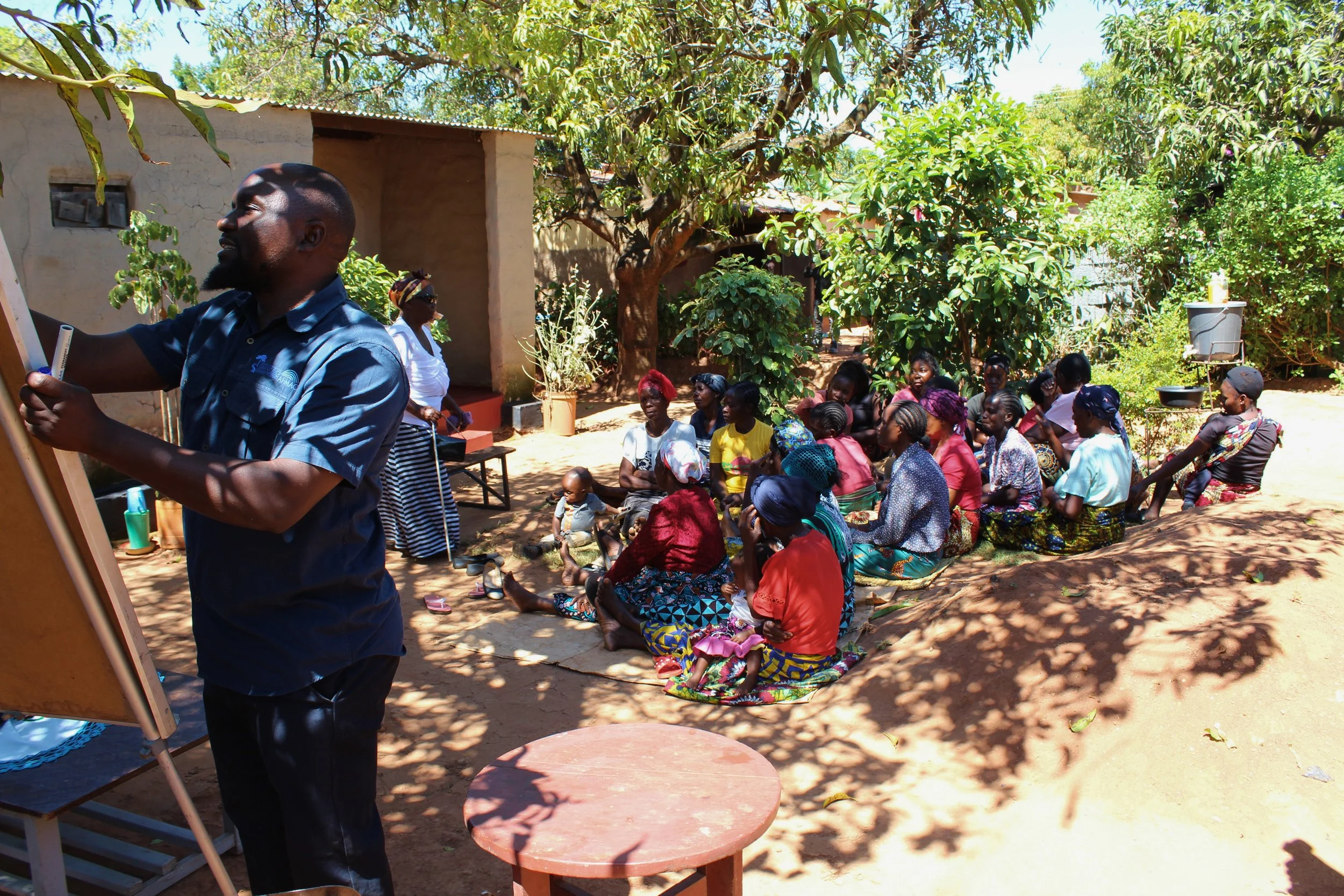 A group of people sitting outdoors under a large tree in a sunny yard, listening to a man in a blue shirt who is standing in front of a whiteboard.