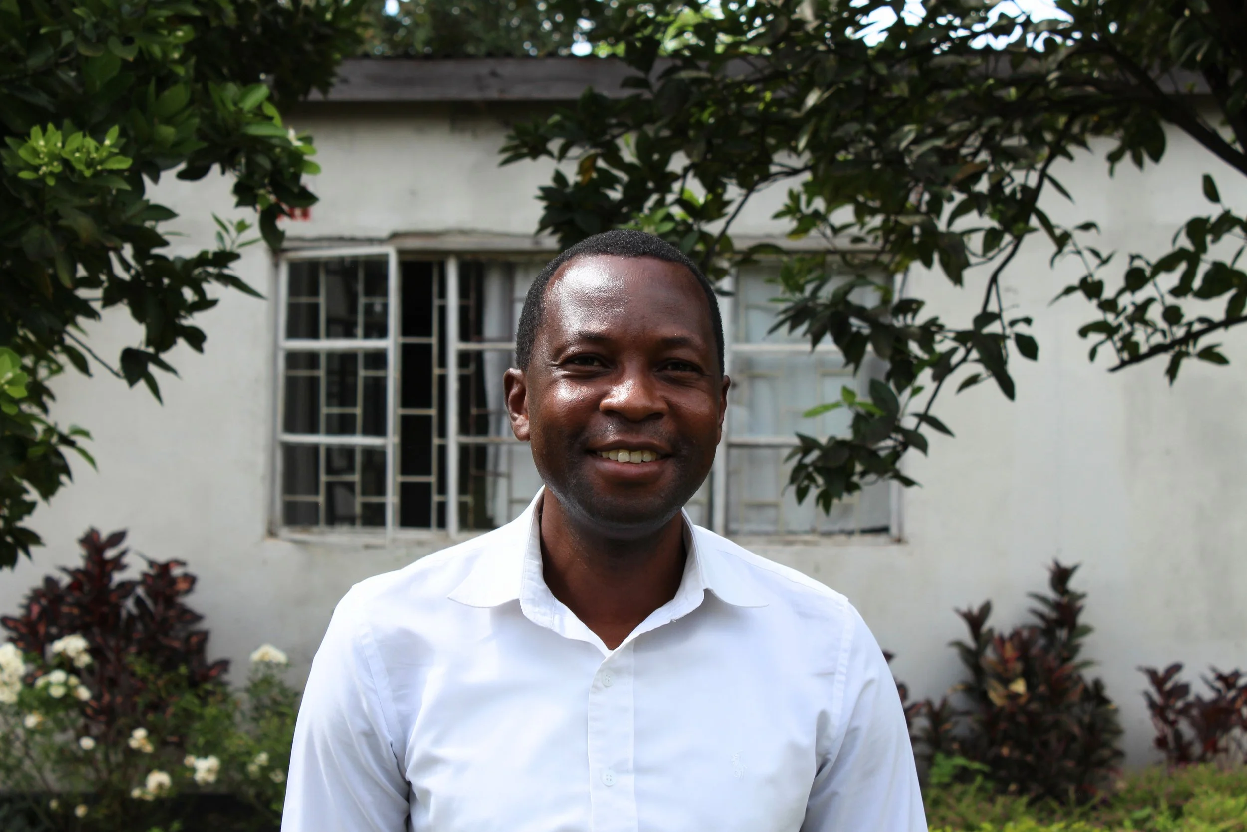 A smiling man in a white shirt standing outdoors in front of a white house with a window, surrounded by green foliage and flowers.