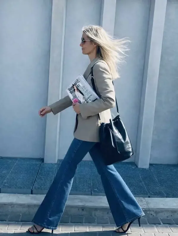 Eine Frau in beige Mantel, blaue Jeans und hohen Schuhen läuft an einer weißen Wand entlang, trägt eine schwarze Tasche und hält Zeitung.