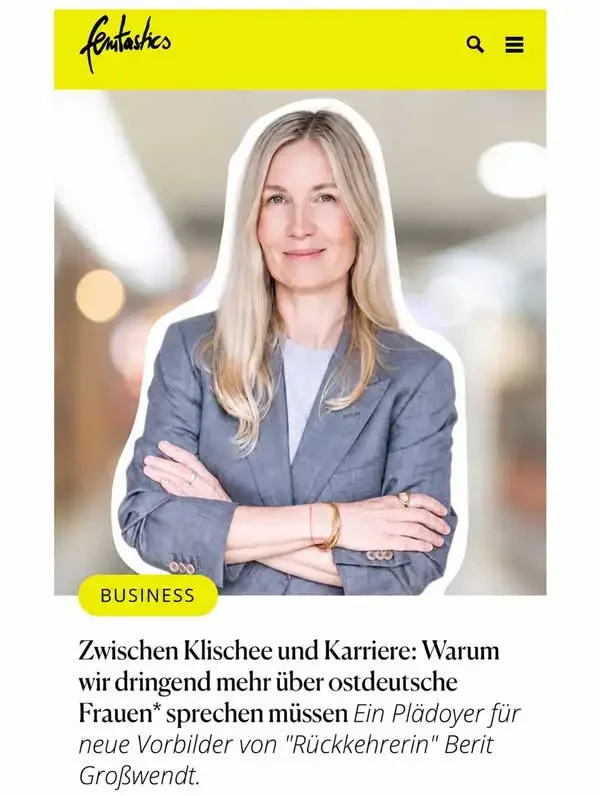 Porträt einer Frau in einem grauen Anzug, die in einem modern ausgeleuchteten Raum steht, mit verschränkten Armen und einem freundlichen Lächeln.