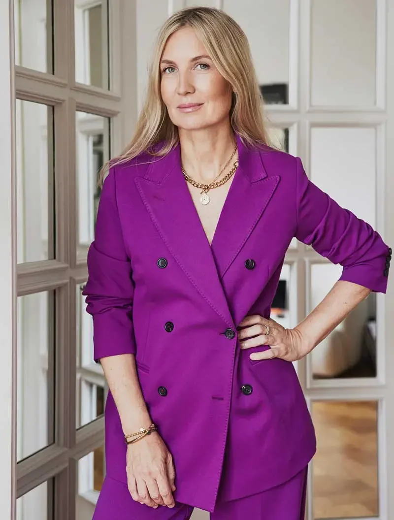 Eine Frau mit langen blonden Haaren trägt einen lila-Blazer und einen Rock oder Hosen. Sie steht vor einer Wand mit quadratischen Fensterrahmen und schaut in die Kamera.