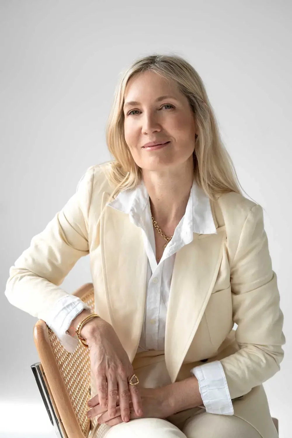 Eine lächelnde Frau mit blonden Haaren, in einem weißen Hemd und cremefarbenem Blazer, sitzt auf einem Stuhl vor neutralem Hintergrund.