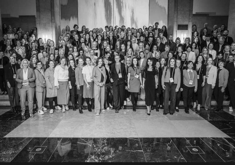 Gruppe von aufgehenden Menschen bei einer Veranstaltung in einem großen Saal, alle lächeln, viele tragen Namenschilder, schwarz-weiß Foto.