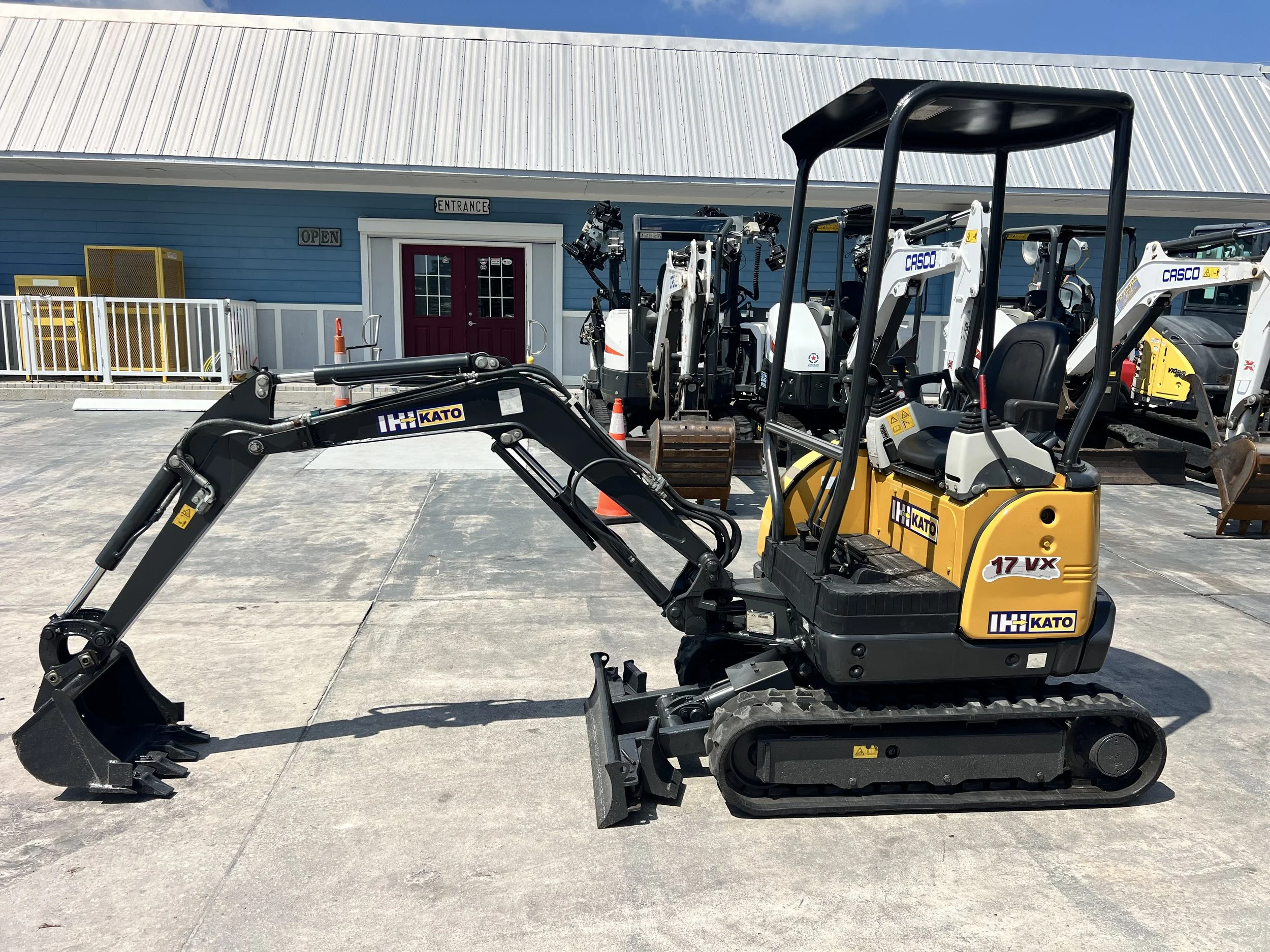 Construction equipment parked outside a building with a blue exterior, grey roof, and double red doors, including compact excavators and machinery with the brand name 'CASCO'.