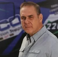 Man with short dark hair and a serious expression standing in front of a race car with the number 22.