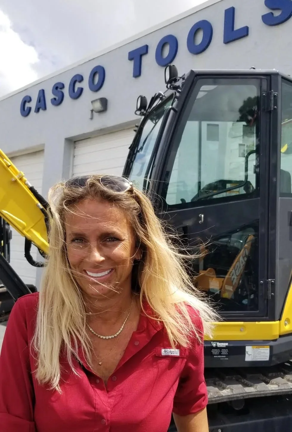 A smiling woman with blonde hair wearing a red shirt and sunglasses on her head, standing in front of a yellow and black construction vehicle outside a building with a sign that reads 'Cascos Tools'.
