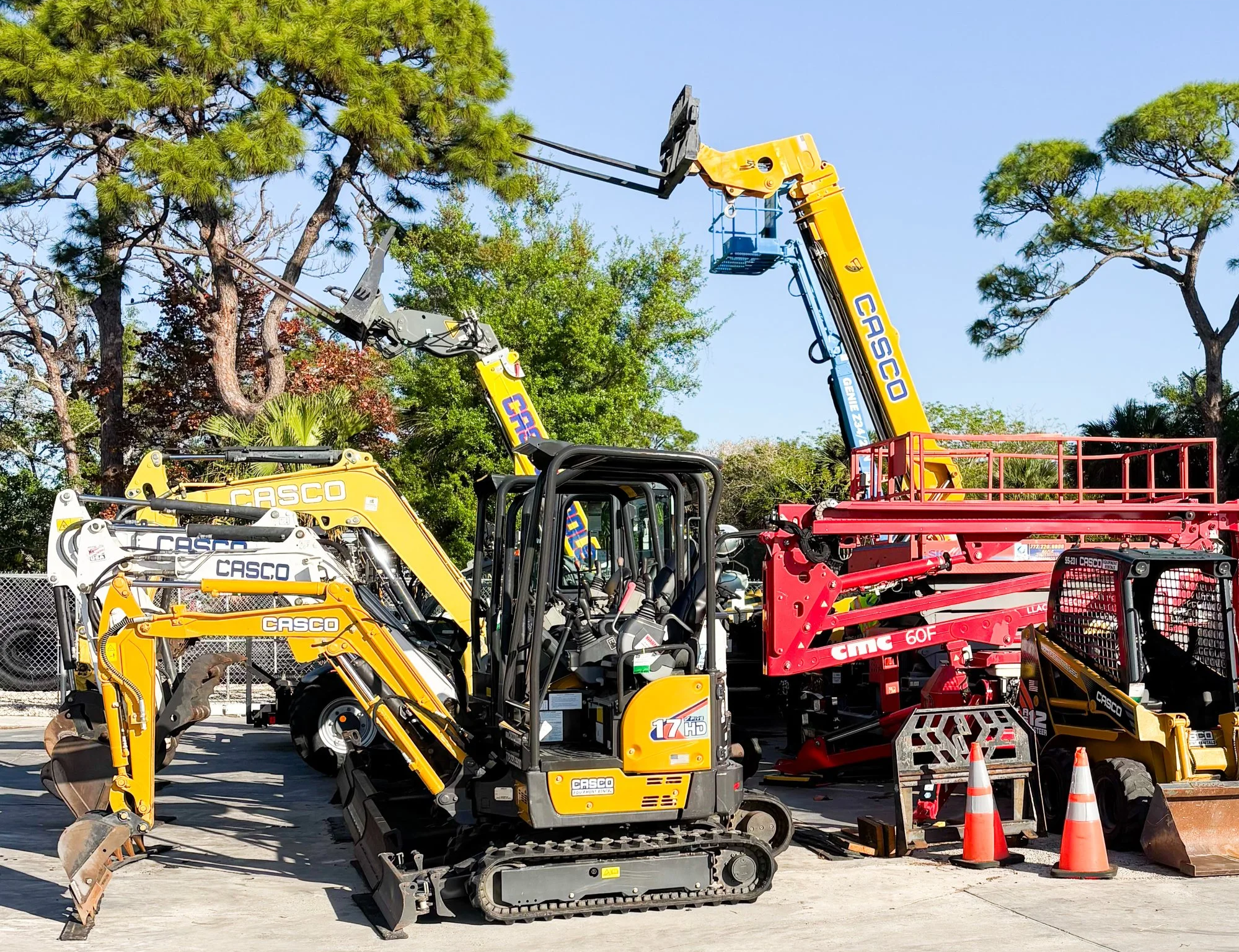 Construction equipment including a yellow mini excavator, a cherry picker, and various machinery and tools are parked outdoors. There are trees and a clear blue sky in the background.